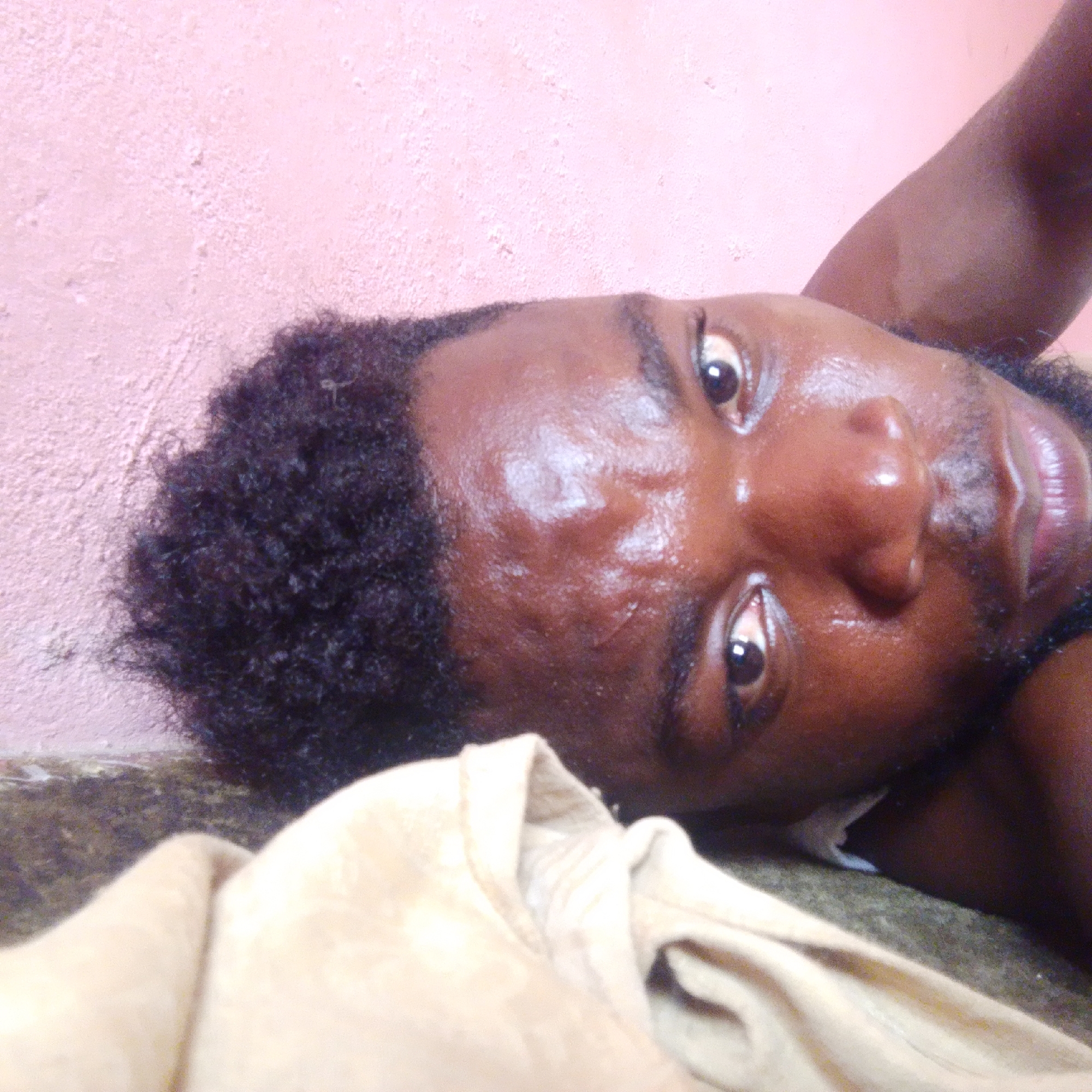 A close-up, high-angle shot depicts a Black male lying down, possibly on a surface, with his head turned towards the camera. His skin appears shiny and possibly sweaty. He has short, curly black hair. A beige or light-colored garment is visible in the foreground, near his neck. The background includes a textured pink wall and a dark, possibly textured surface beneath his head. Location: Unknown, Unknown.