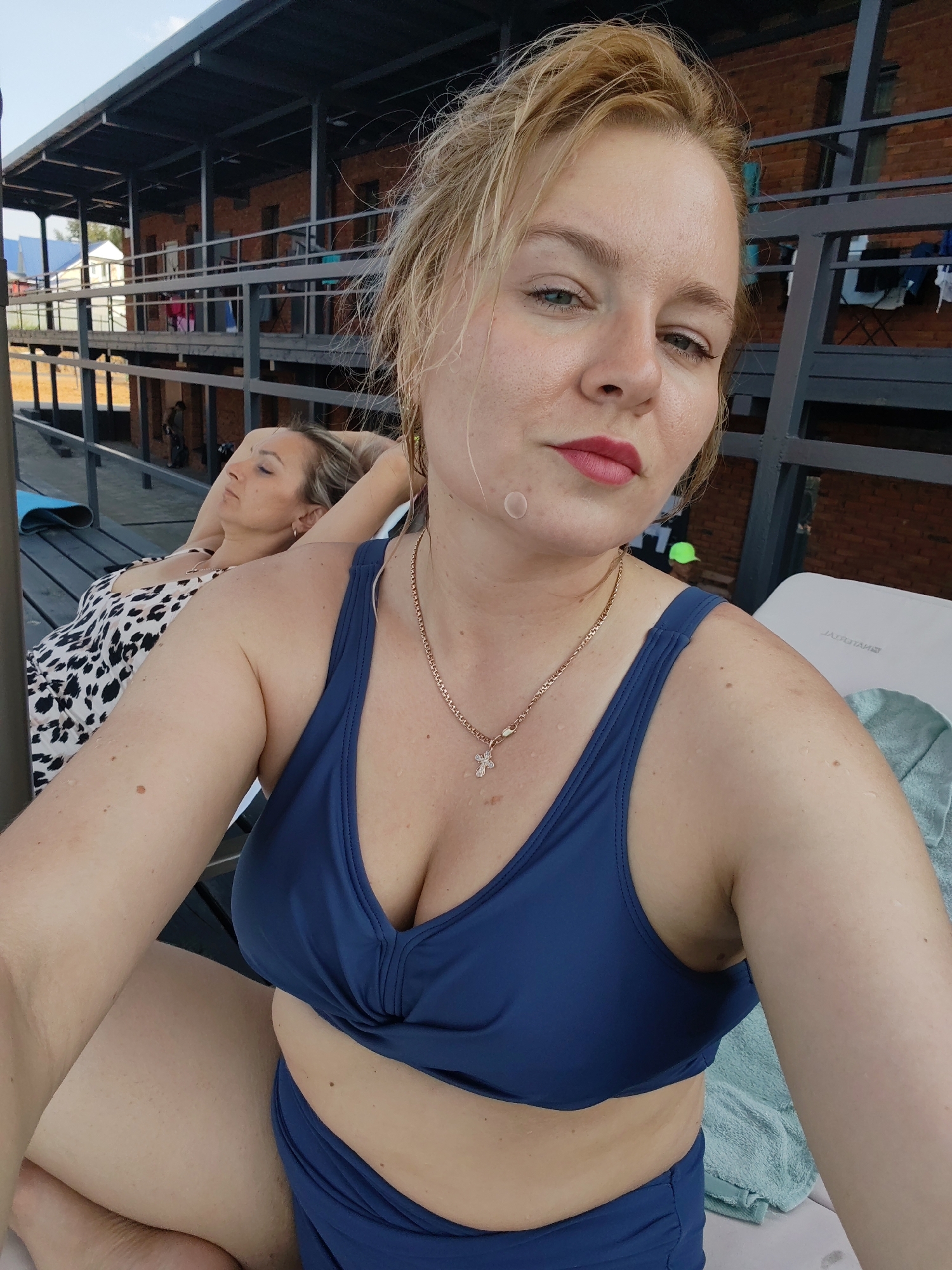 A woman with blonde hair and makeup is taking a selfie with a blue bikini. Another woman is lying on a lounge chair behind her, wearing a black and white patterned dress. They appear to be outdoors near a building with multiple levels and a metal roof. The location is identified as Pirogovo, Russia.