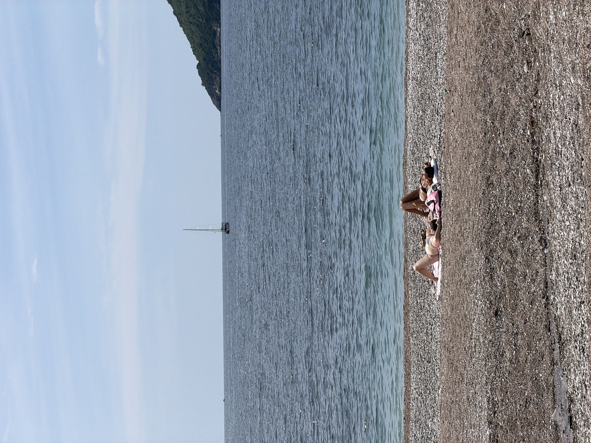 The image depicts a scenic beach view in Budva, Montenegro. Two people are lying on a beach towel on the pebble-covered beach. The sea is calm and reflects the sunlight. In the distance, a sailboat is visible. The sky is blue. A lush, green, mountainous coastline forms part of the backdrop.