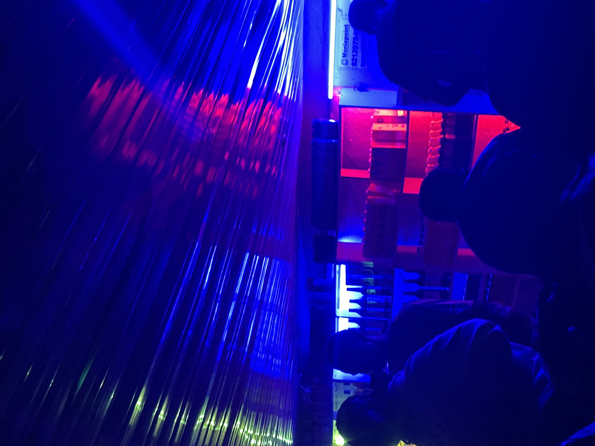 The image depicts an indoor scene, possibly a bar or club, illuminated by vibrant blue and red lights. In the foreground, silhouettes of several individuals are visible, facing away from the camera. The individuals appear to be standing or sitting. The background reveals shelves stocked with bottles, some of which are illuminated by the colored lighting. A sign with text, including "Moniepoint" and a numerical code, is partially visible on a white surface in the mid-ground. The ceiling is covered with what appears to be corrugated material, reflecting the light and creating a striped effect. The overall atmosphere is dark and dimly lit, with the colored lights creating a dramatic effect. The location is identified as Wumat, Nigeria.