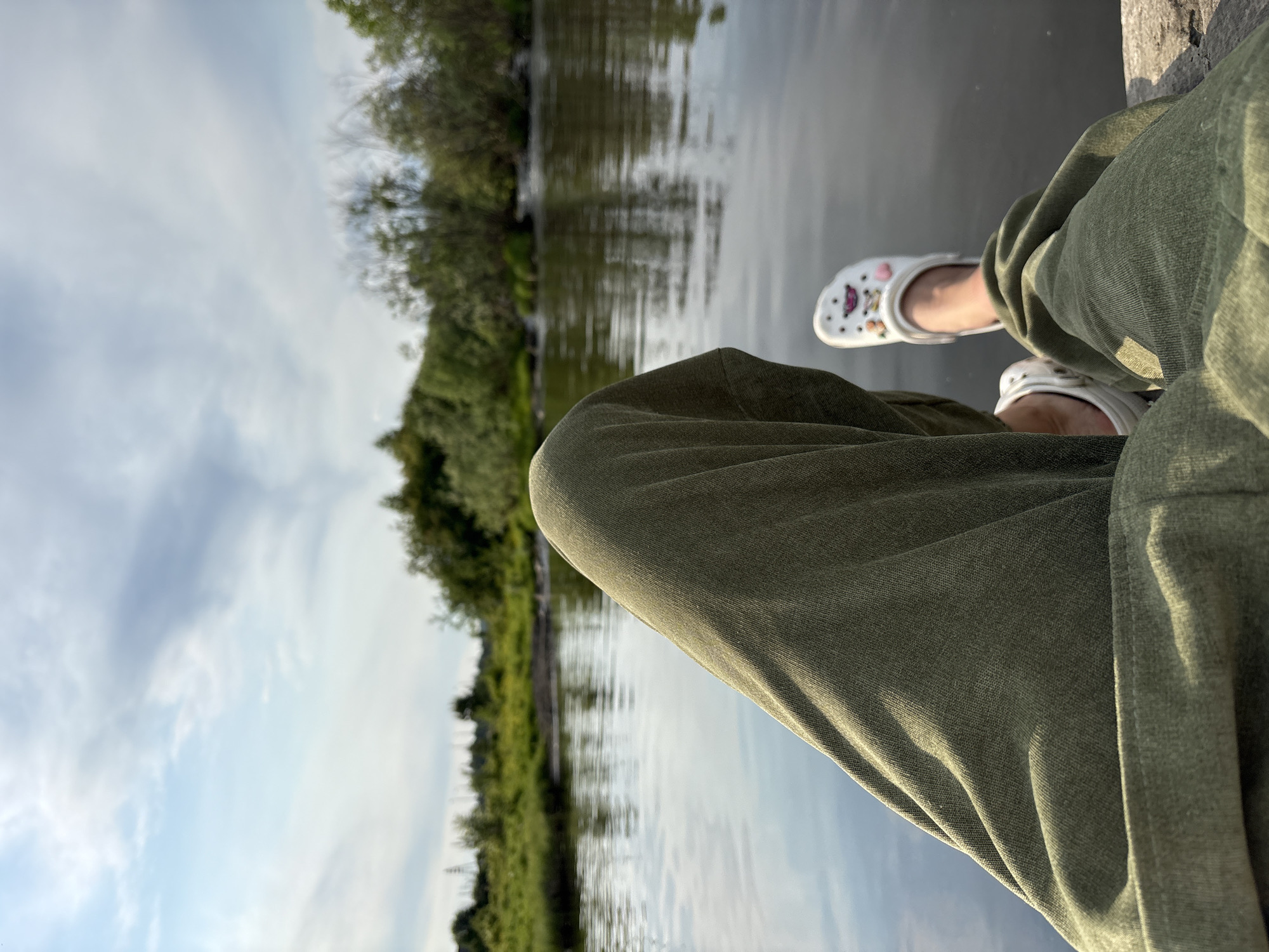 The image shows a close-up perspective of a person's legs and feet, clad in olive green pants and white Crocs, resting on a stone ledge by a body of water. The water's surface reflects the sky and the surrounding lush green trees and vegetation, creating a serene natural landscape. The sky is partly cloudy, with patches of blue visible through the white clouds. The overall scene suggests a moment of relaxation in an outdoor setting, possibly near the city of Nevon, Russia, given the natural environment.