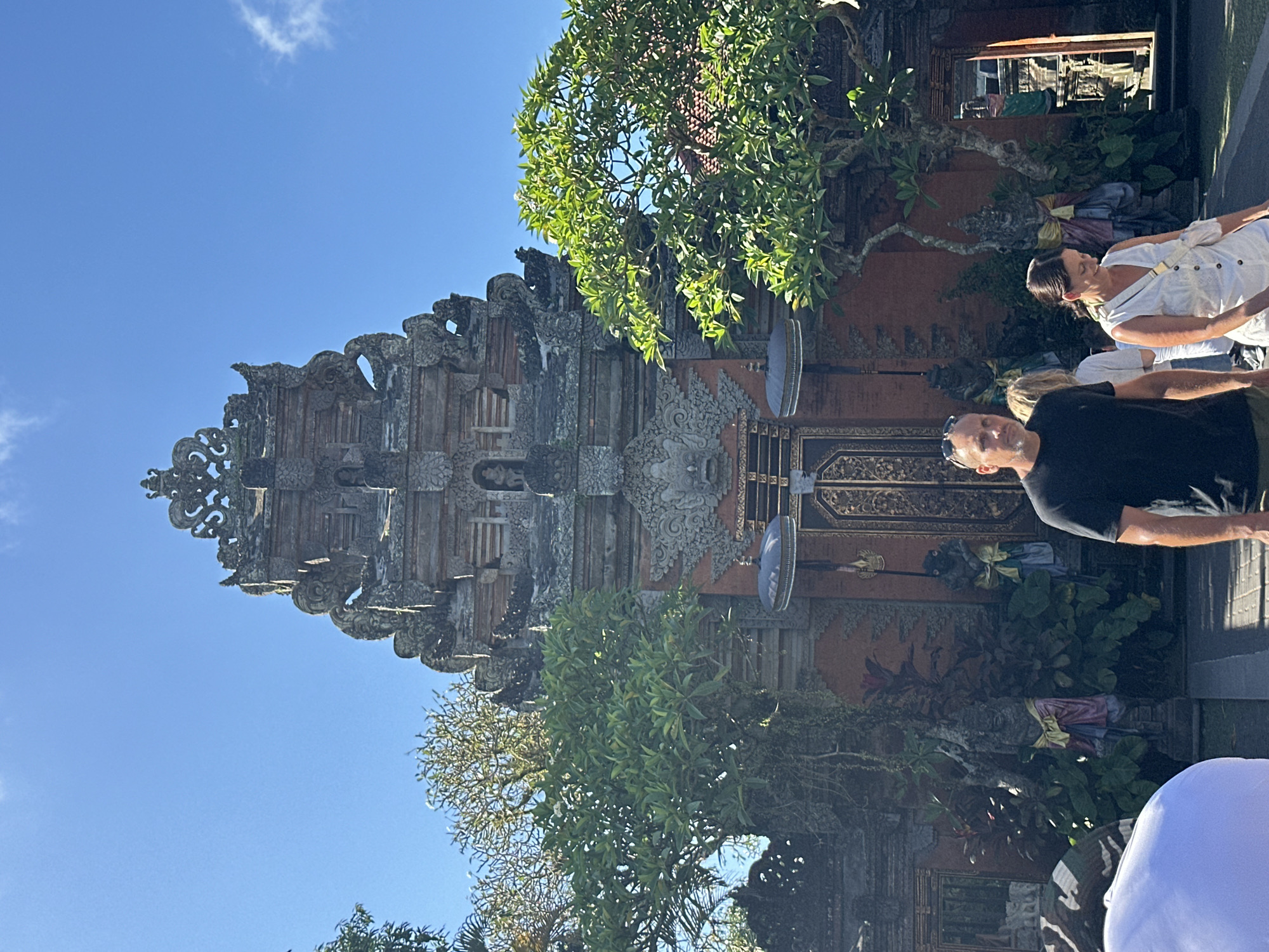 In Ubud District, Indonesia, a man in a black t-shirt and shorts walks towards the camera, while a woman in a white dress walks past him. Behind them is a traditional Balinese temple entrance, featuring intricate stone carvings, ornate doors, and two large trees. The sky is clear and blue.