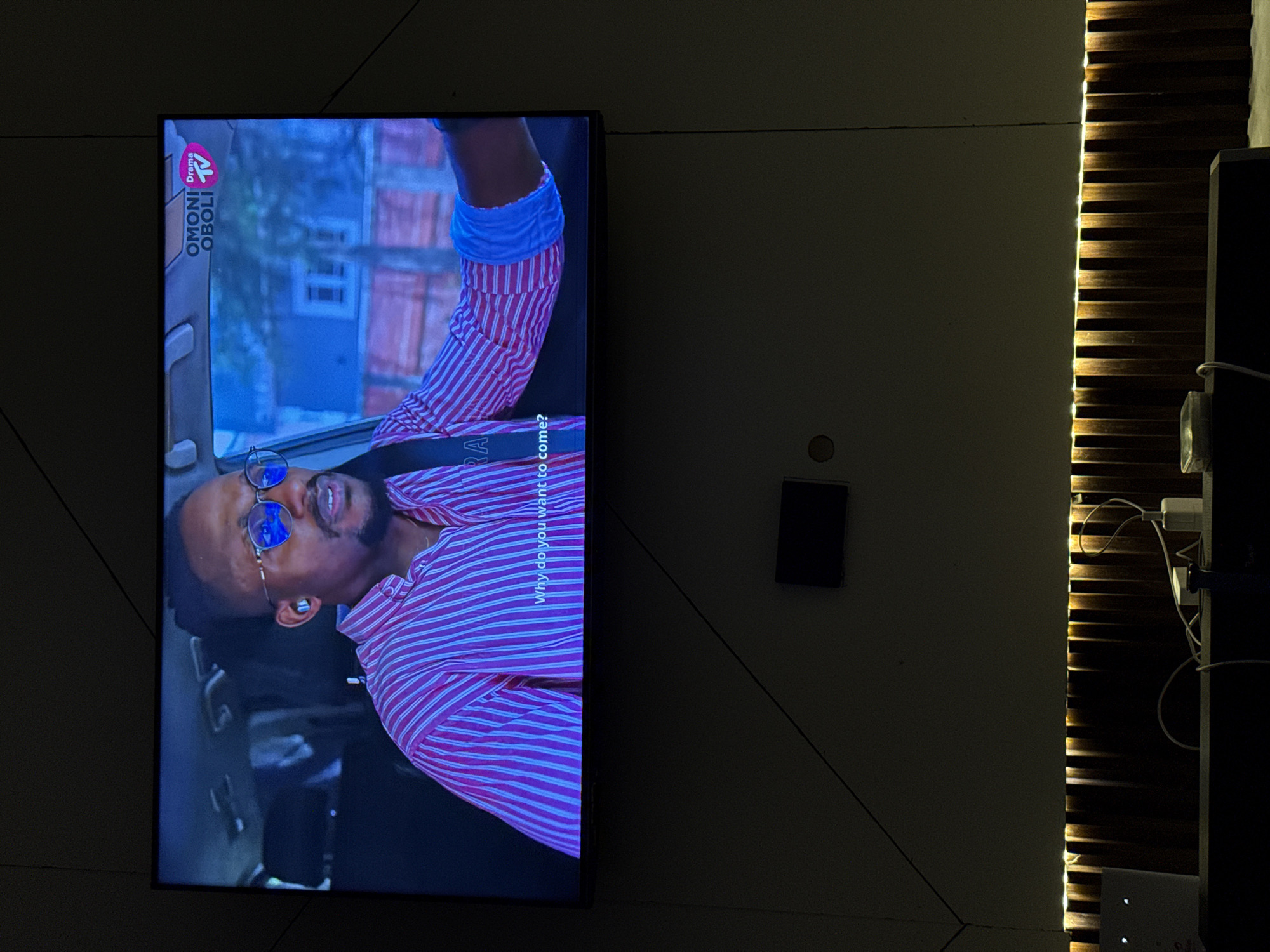 The image shows a television mounted on a wall in what appears to be an interior space. The TV screen displays a scene of a dark-skinned man wearing sunglasses and a pink and white striped shirt, potentially in a car. The text "Why do you want to come?" is displayed on the screen. To the right of the TV, there's a decorative wooden feature with integrated lighting. Below the wooden feature, there are visible electrical outlets with plugs and cables. The image does not provide enough information to locate it in Iddo Tudunwada, Nigeria.