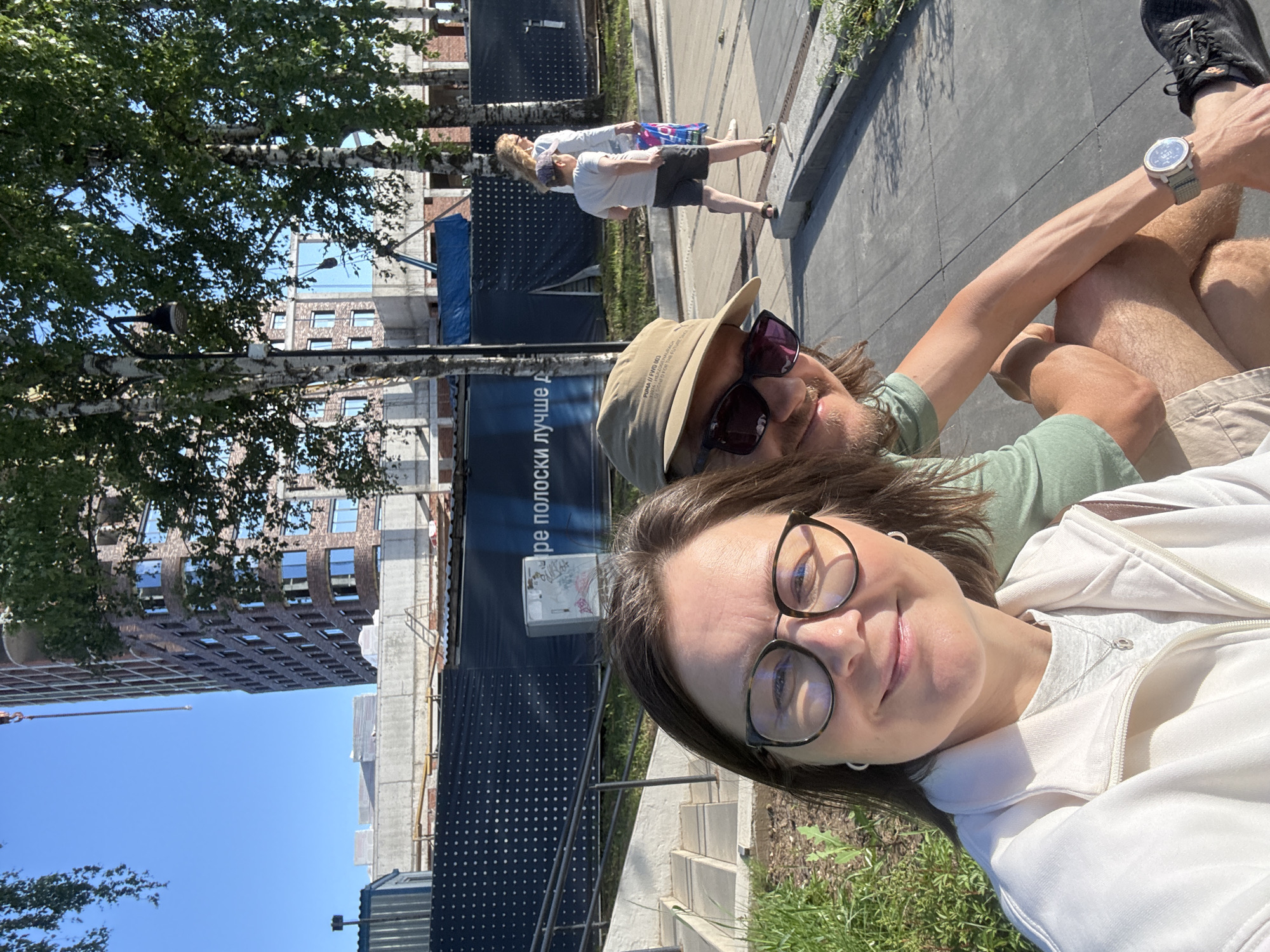 In Izhevsk, Russia, a selfie is captured in a sun-drenched urban environment, likely near a construction site. The foreground features a couple, with the woman on the left wearing glasses and a white jacket, and the man on the right sporting a tan baseball cap and sunglasses. They are both smiling at the camera. The background reveals a multi-story building under construction, with scaffolding and cranes visible. Trees with green foliage are interspersed throughout the scene, casting shadows on the paved ground. Further in the background, two other individuals, a woman and a child, are walking away from the camera. A dark blue banner with white text is stretched across part of the construction site. The overall atmosphere is casual and bright, suggesting a pleasant day outdoors.