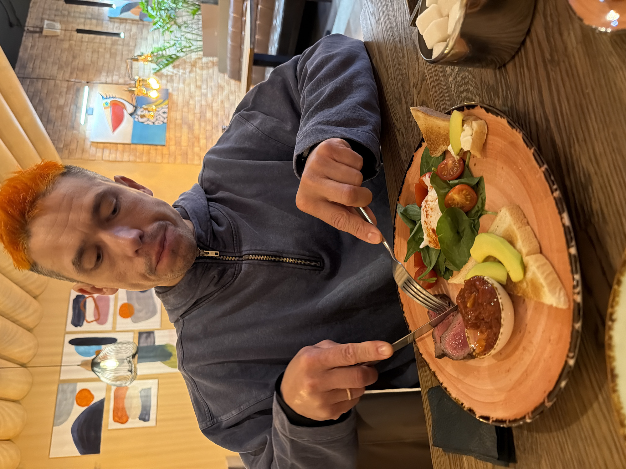 A person is eating a meal in a restaurant. The person, with close-cropped orange hair, is wearing a dark blue zip-up jacket. They are using a knife and fork to cut into a dish served on a decorative orange plate. The plate contains steak, a white, round food item (possibly cheese), avocado slices, cherry tomatoes, greens, and small slices of toast. A container of small, white cubes sits nearby on the wooden table.
The restaurant has distinctive wall decorations, including abstract paintings and a neon sign.
The prompt mentioned the city of Chaikovsky, Russia, but there is no way to confirm this information based on visual evidence alone.