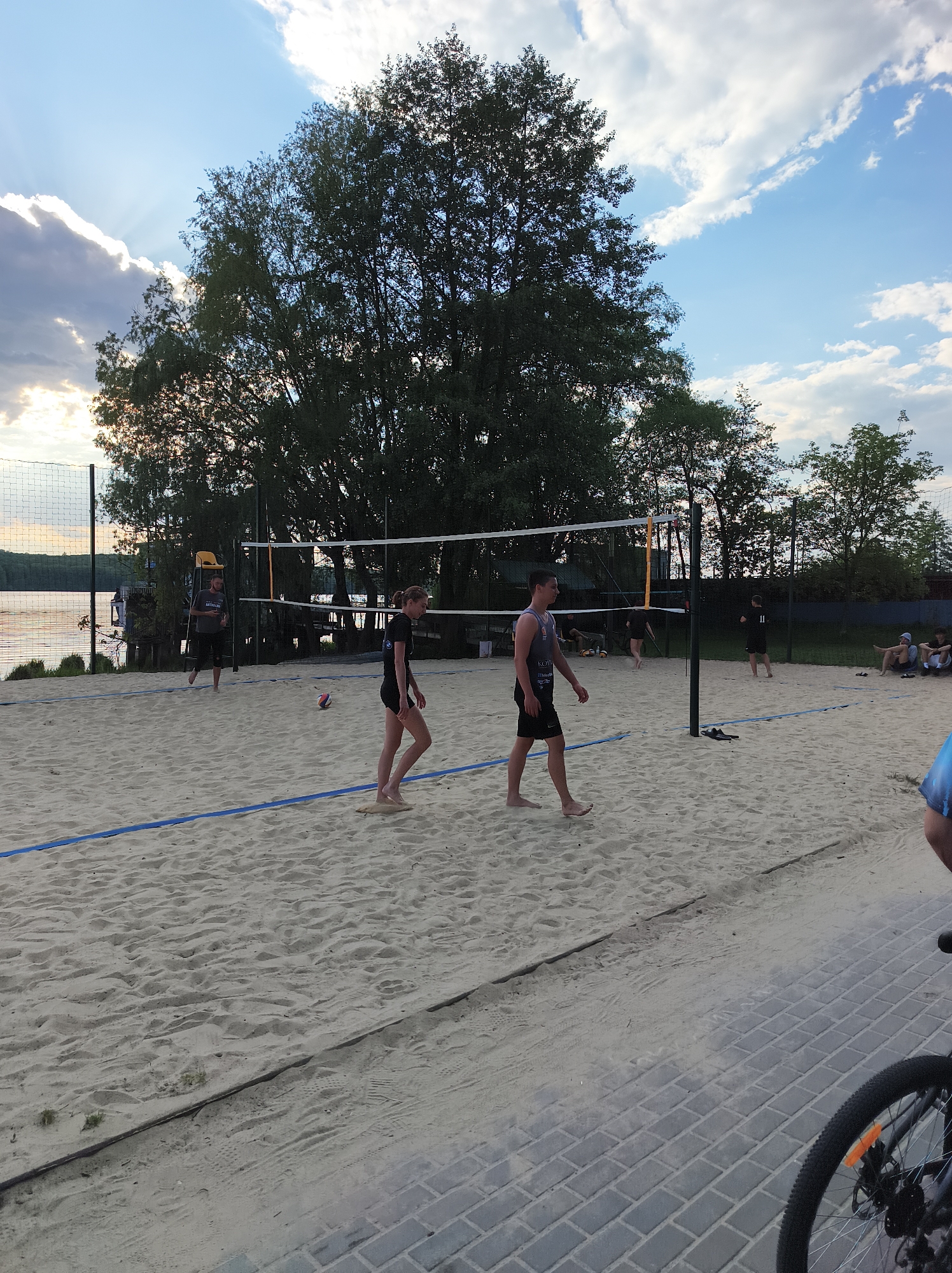 The image captures a beach volleyball scene in Ternopil, Ukraine. A sandy court is visible, marked with blue lines, with a volleyball net stretched across the center. Several people are on the court, including a young woman and a young man walking barefoot near the net. Other individuals are scattered around the court, some playing or observing the game, and others sitting. In the background, a lake is visible behind the volleyball net, with trees lining the shore. The sky is a mix of blue and clouds, suggesting late afternoon or early evening light. In the lower right corner of the image, part of a bicycle wheel is visible. A paved area separates the sand from the viewer.