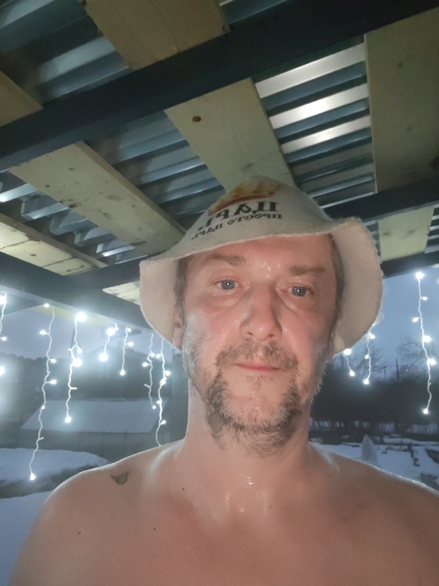 A middle-aged man with wet skin and wearing a sauna hat stands outdoors in what appears to be a snowy environment. He is shirtless. The environment features a structure with exposed wooden beams and metal panels overhead. Fairy lights hang in the background, partially obscured by snow and trees. The scene is identified as being located in Chaikovsky, Russia.
