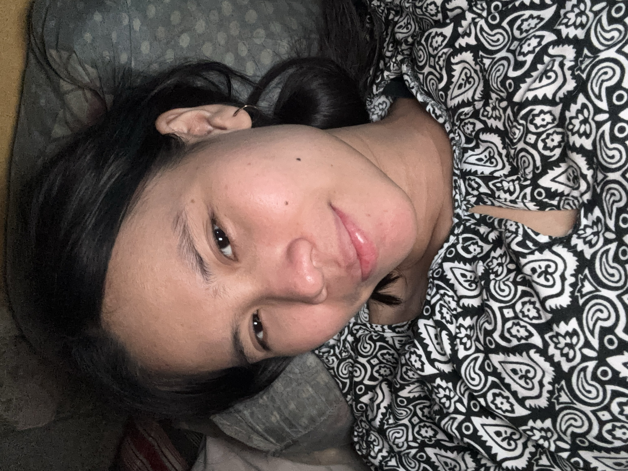 A close-up, selfie-style shot features a young Indonesian woman lying down on a bed. She has dark hair and appears to be looking directly at the camera with a slight smile. She is wearing a black and white patterned top. The background is blurred, but appears to be a bedroom setting with pillows and bedding. The image is likely from East Jakarta, Indonesia, given the provided context.