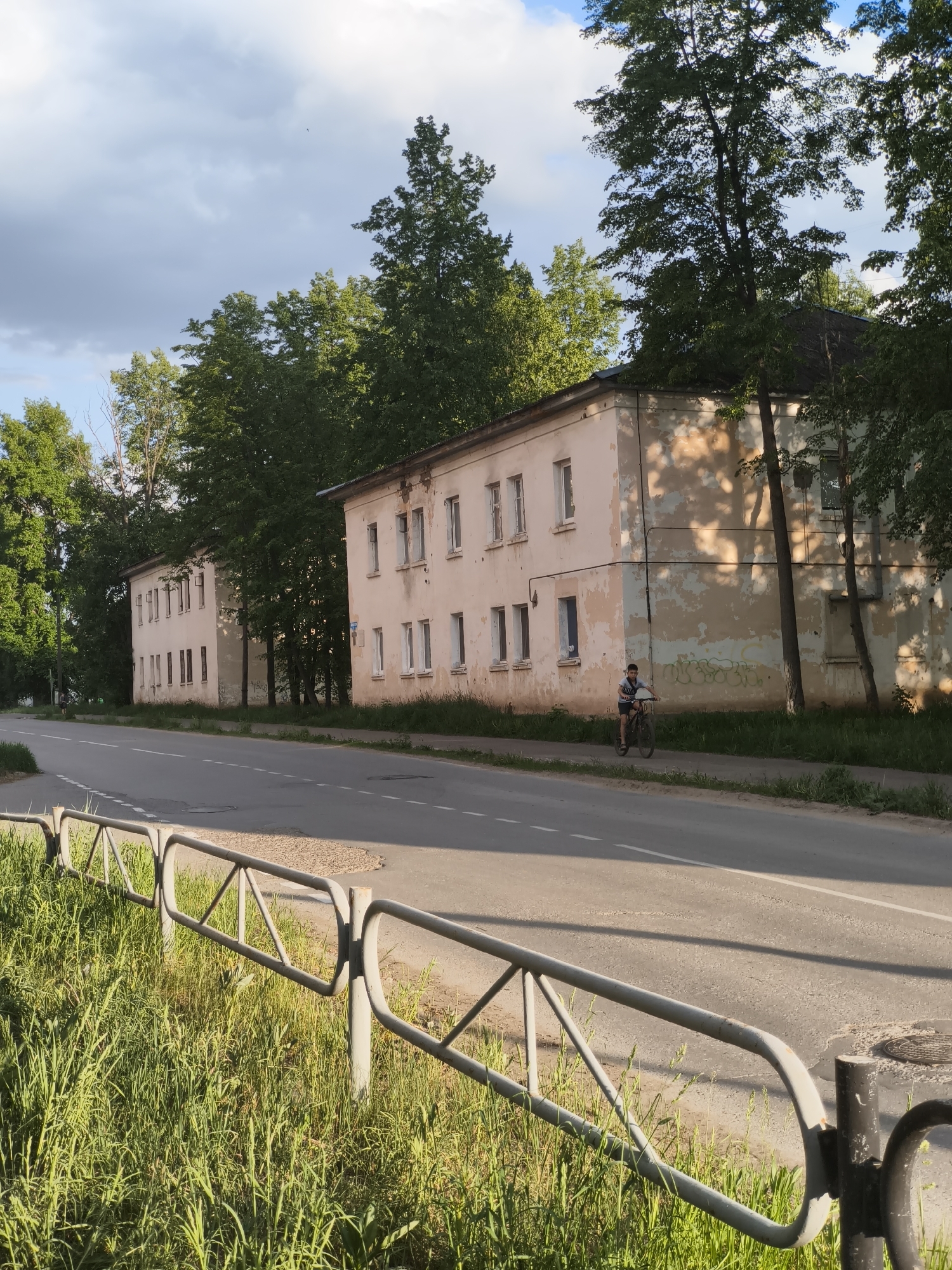 The image depicts a street scene in what appears to be a residential area in Chaikovsky, Russia. Two identical, dilapidated, two-story buildings with peeling pale pink paint are aligned along the roadside. The buildings have rectangular windows with white frames. A young boy is riding a bicycle on the sidewalk next to the buildings. The street is paved with a white centerline marking. A metal guardrail runs along the edge of the road, separating it from a grassy area with tall green grass. The sky is partially cloudy. Trees line the buildings, obscuring parts of them. The overall scene is mundane, showing a quiet, possibly aging neighborhood.