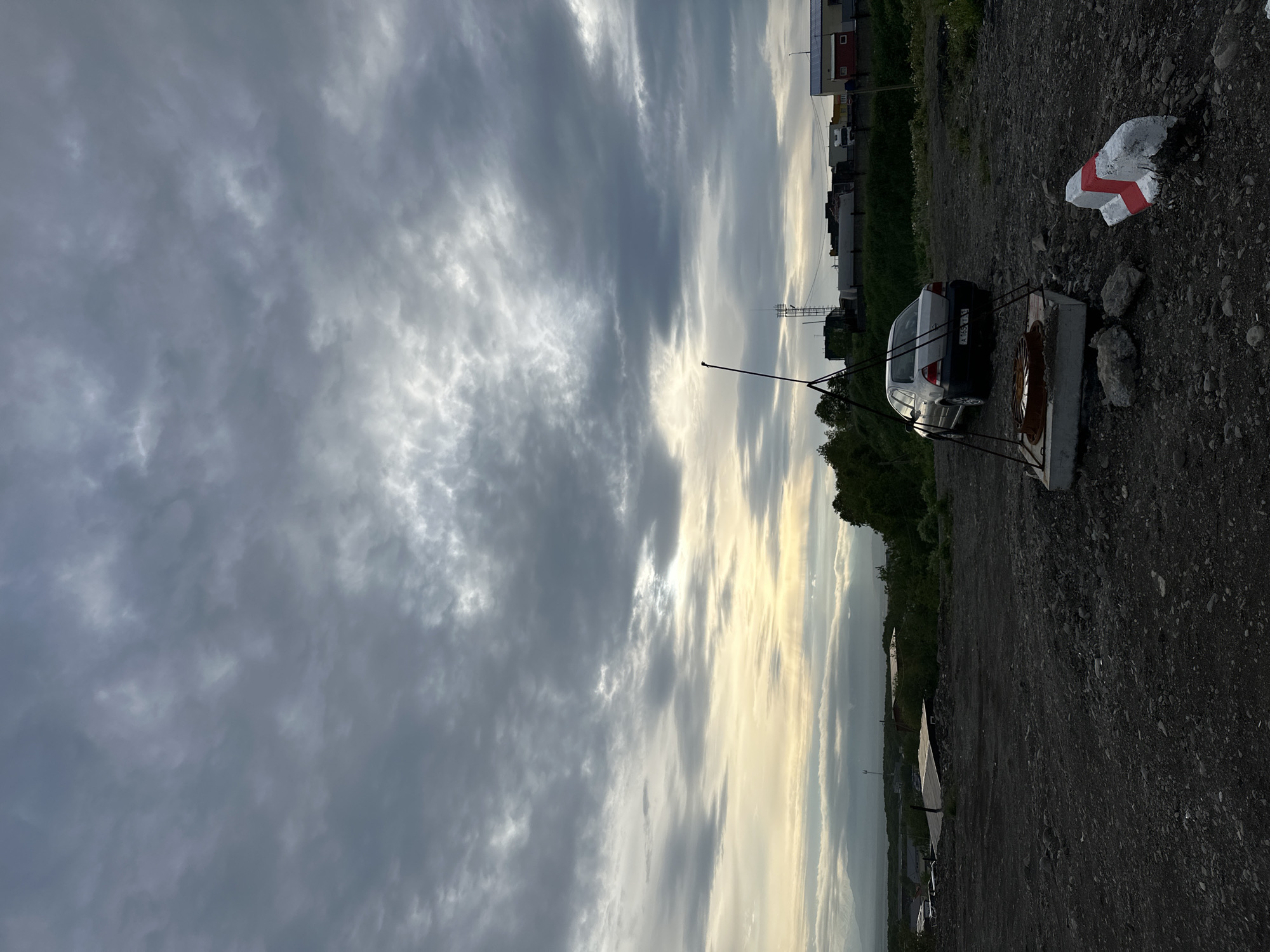 The image depicts an overcast sky above a rural or industrial outskirts area, likely in Petropavlovsk-Kamchatsky, Russia. The foreground consists of a gravelly, uneven ground with scattered stones and debris. In the mid-ground, a silver sedan is parked. Near the car, a concrete structure with a metal grate is visible, supporting what appears to be an antenna or a measuring device. A red and white concrete barrier block lies on the ground to the right of the structure. In the background, a line of low-rise buildings and industrial structures is present, along with some green vegetation and trees. A distant mountain range is faintly visible through the hazy atmosphere. The overall impression is one of an undeveloped or utilitarian landscape under a dramatic, cloudy sky.