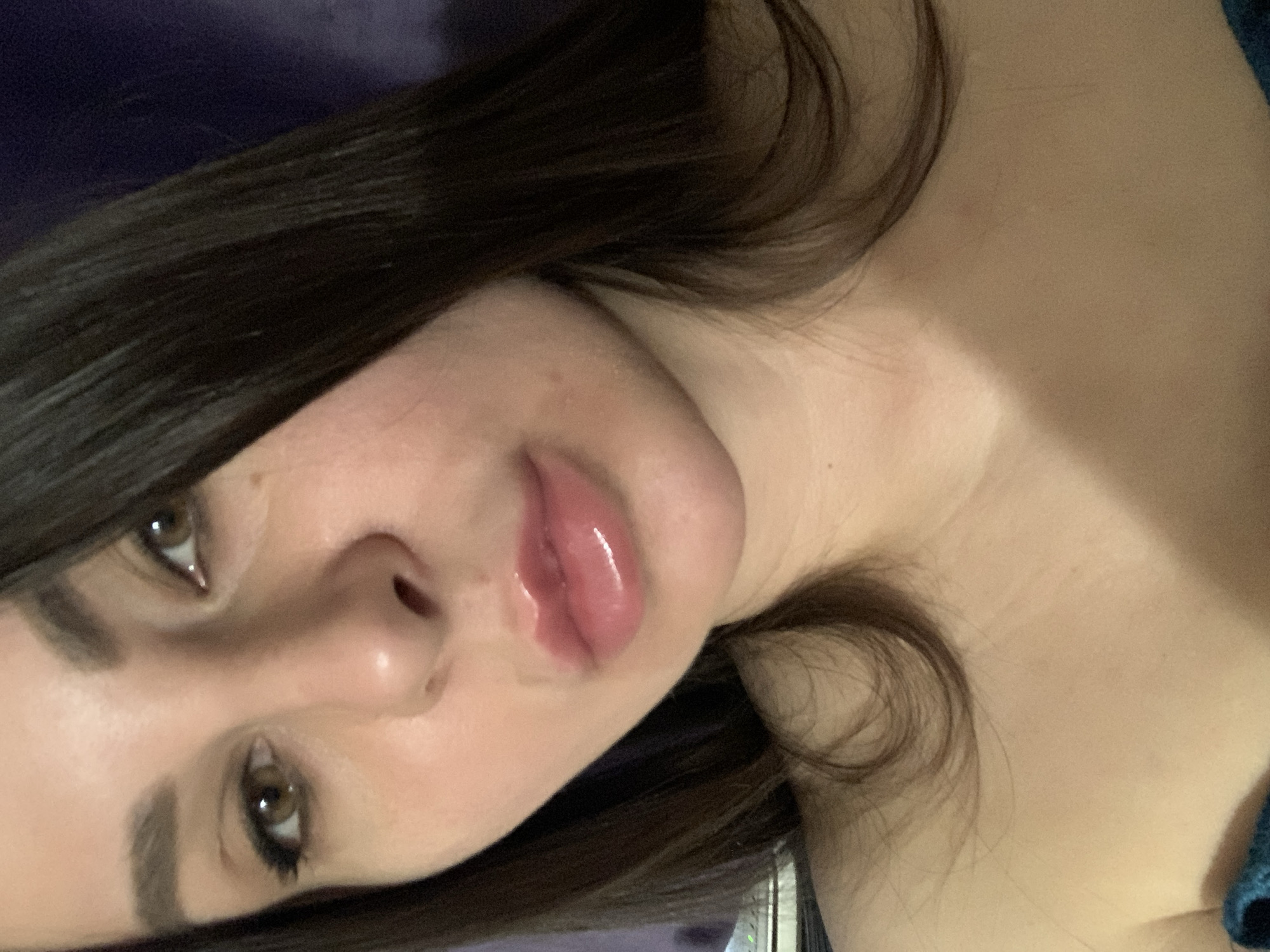 A close-up, head-and-shoulders shot shows a young woman with dark brown hair and brown eyes. She is looking directly at the camera. Her lips are full and glossy, with a pinkish-nude lip gloss. The lighting is soft, and the background is a blurred, dark purple. She is wearing a dark blue top. The image appears to be a selfie, taken indoors.