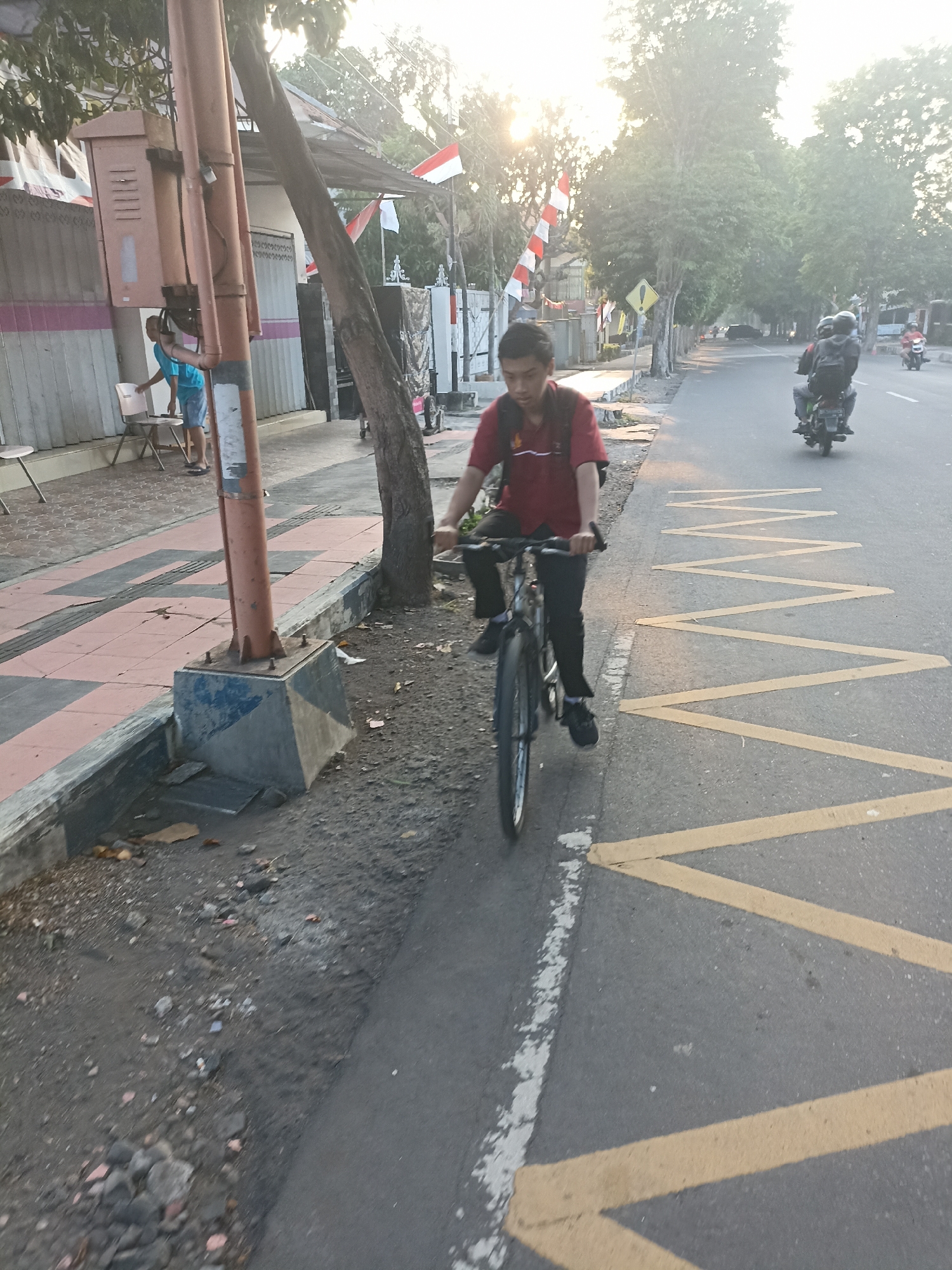 In Mojopanggung, Indonesia, on a paved road with yellow zigzag markings and a white dashed line, a young man wearing a red polo shirt and black pants is riding a bicycle. He is positioned on the right side of the road, traveling towards the viewer. A tree trunk stands to the left of the road, adjacent to a sidewalk paved with reddish-orange and gray tiles. A lamppost with an orange pole is situated near the tree. In the background, two individuals on a motorcycle are also on the road, heading away from the viewer. Further in the distance, more trees and buildings are visible, with Indonesian flags displayed. The scene is illuminated by the setting sun, casting a warm glow.