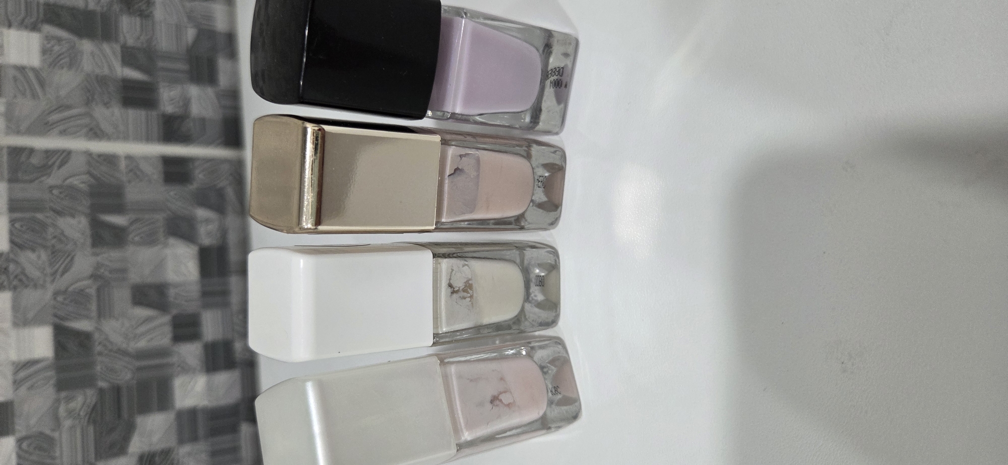 The specific city of Lipetsk, Russia, cannot be determined from the visual evidence. The scene depicts four rectangular glass bottles of nail polish, arranged in a horizontal line on a smooth, light-colored surface. The background features a tiled wall with a repetitive gray and white geometric pattern. No people or actions are present.
From left to right:
1. The leftmost bottle has a translucent, off-white cap and contains a pale pink nail polish. The polish level is low, and dried remnants are visible on the interior glass walls.
2. The second bottle has a solid white, opaque cap and contains an off-white or cream-colored nail polish. The liquid level is low, with pronounced dried and discolored polish clinging to the interior.
3. The third bottle has a reflective gold-colored cap and contains a pale peach-toned nail polish. The liquid level is low, and dried polish residue is visible inside.
4. The rightmost bottle has a solid black, opaque cap and contains a light lavender or lilac nail polish. This bottle's polish level appears comparatively higher than the others.
The light-colored surface beneath the bottles exhibits subtle reflections and minor surface imperfections.