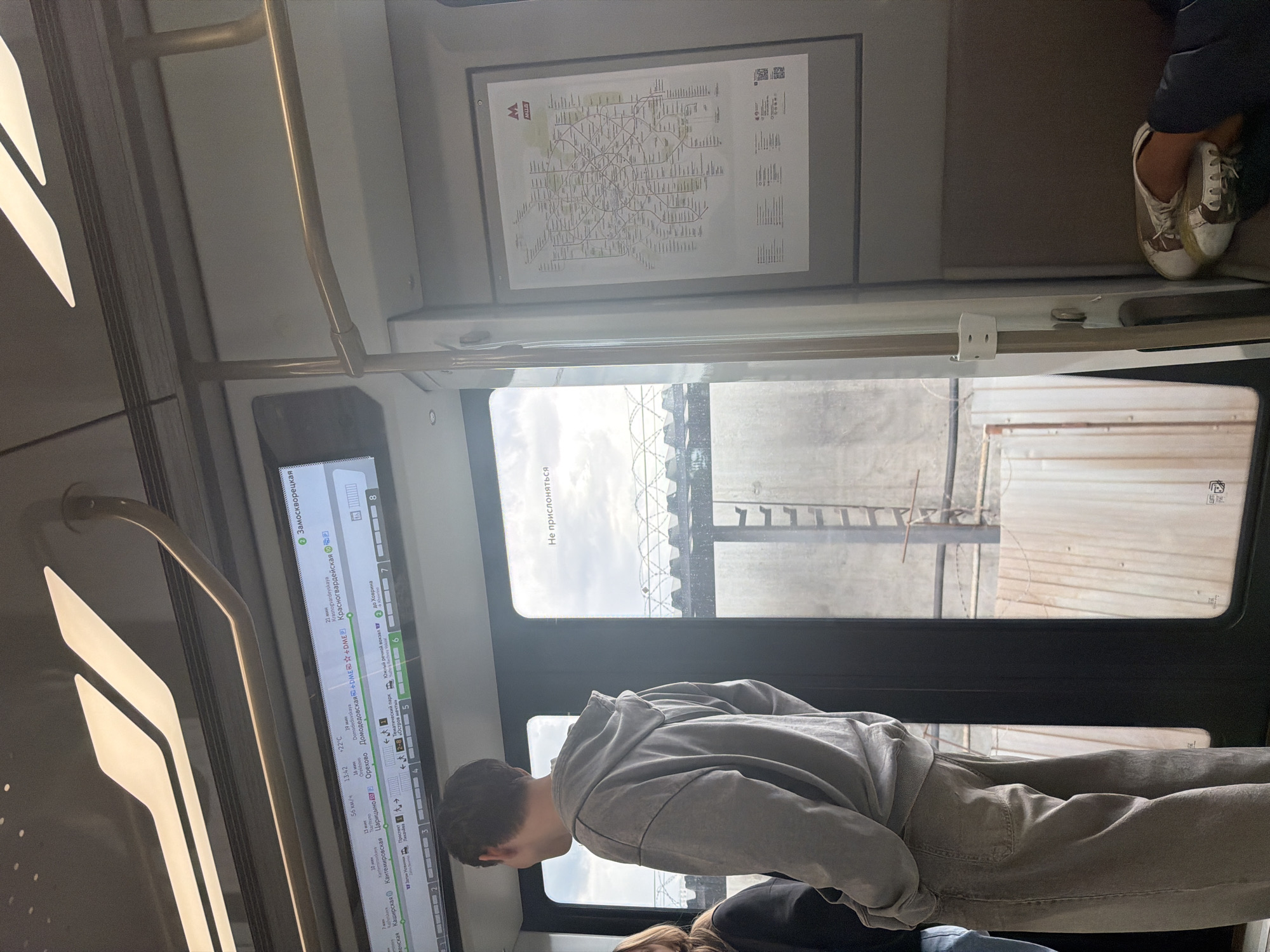 The image shows the interior of a subway train. In the foreground, a young person wearing a gray hoodie and loose-fitting pants stands facing away from the camera. Their hands are in their pockets, and they are looking out of the train door. To the left, a portion of another person wearing dark clothing is visible.
A digital display above the train door shows route information, including station names, times, and temperatures. The display shows "Замоскворецкая" (Zamoskvoretskaya) at the top right.
To the right of the door, a subway map of Moscow is displayed on the wall. The map features the familiar red "M" logo of the Moscow Metro. Outside the train door, a view of what appears to be an industrial area or train yard is visible through a large window. Barbed wire fences and some structures can be seen.
The lighting in the train is a mix of interior fluorescent lights and natural light from the windows. The overall scene is one of everyday transit within a large city.