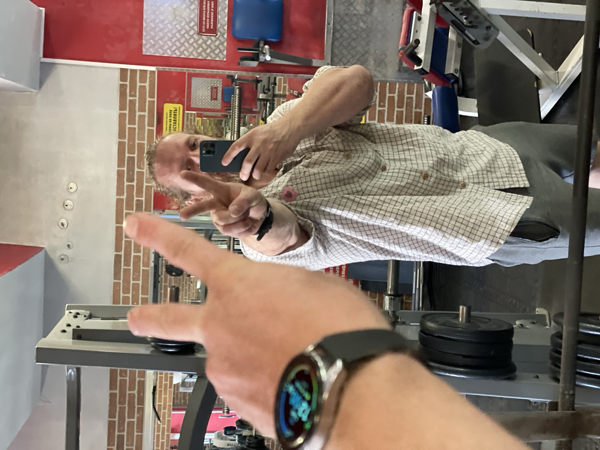 A man is taking a mirror selfie in a gym. He is wearing a plaid shirt and grey sweatpants, and has a watch on his left wrist. He is holding his phone in his right hand and making a peace sign with his left hand. The gym appears to be in a Soviet-era building, evidenced by the brick walls and older gym equipment. The image does not contain enough information to determine the exact location in Saint Petersburg, Russia, or the specific gym.