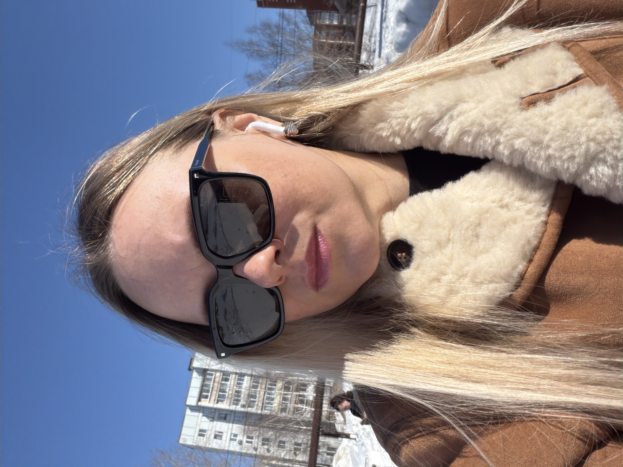 The image displays a close-up outdoor portrait of a female in Votkinsk, Russia, under a clear blue sky. The subject has long, straight, blonde hair and wears large black square-framed sunglasses. A white wireless earbud is present in her right ear, with a small, possibly jeweled, earring visible beneath it. Her lips are a light pink. She is dressed in a brown suede-like coat featuring a prominent cream-colored shearling collar and lapels, with a dark garment visible underneath. A single dark button is visible on the coat.
The background reveals a snow-covered urban environment, indicative of winter or early spring. A multi-story, light-colored apartment building, characteristic of Soviet-era architecture, is visible in the left mid-ground. Bare trees are scattered throughout the scene, and faint wire-like structures are discernible. A blurry, undefined individual is partially visible in the distant left background, behind another structure. A reddish-brown building is also present in the far right background.