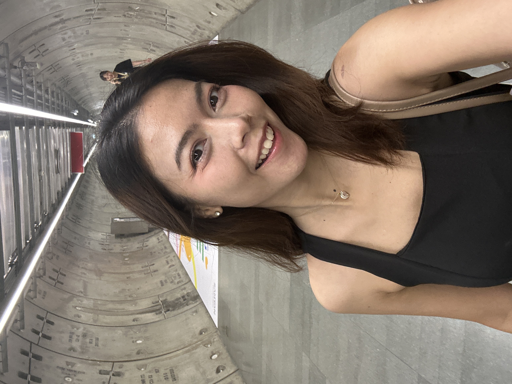 A medium-shot selfie shows a woman with brown hair and fair skin smiling broadly in a concrete tunnel. She is wearing a black sleeveless top and a heart-shaped pendant necklace. In the background, another woman in a black shirt is visible in the distance, talking on a phone. The tunnel walls are made of concrete segments with visible markings, and overhead lighting illuminates the space. A colorful, abstract design is visible on a wall in the mid-ground. This appears to be a transportation tunnel, possibly in Bangkok, Thailand, given the concrete architecture and the presence of public art installations often found in the city's transit systems.