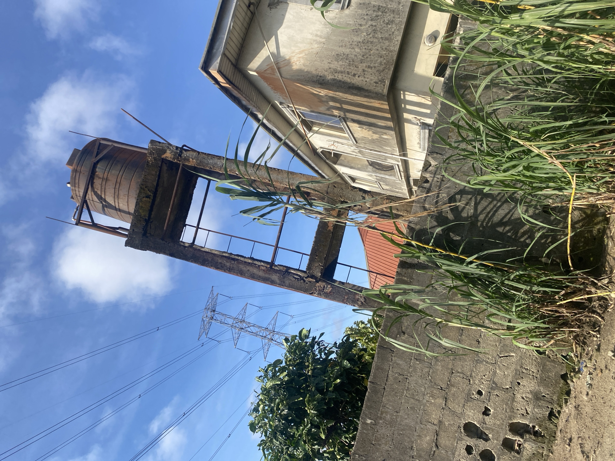 The image depicts a view from the ground level in Ado, Nigeria, looking upwards. In the foreground, tall green stalks of sugarcane grow along a rough, grey concrete wall. Behind the wall, a weathered, two-story building with multiple windows and visible water stains on its exterior is present. To the left, a lush green tree with visible fruit can be seen. Dominating the upper portion of the image are high-voltage power lines supported by a large metal pylon. In the middle ground, an old, rusty water tank rests precariously on a decaying concrete structure that appears to be an elevated water tower, with a ladder-like metal frame attached to it. The sky is bright blue with scattered white clouds.