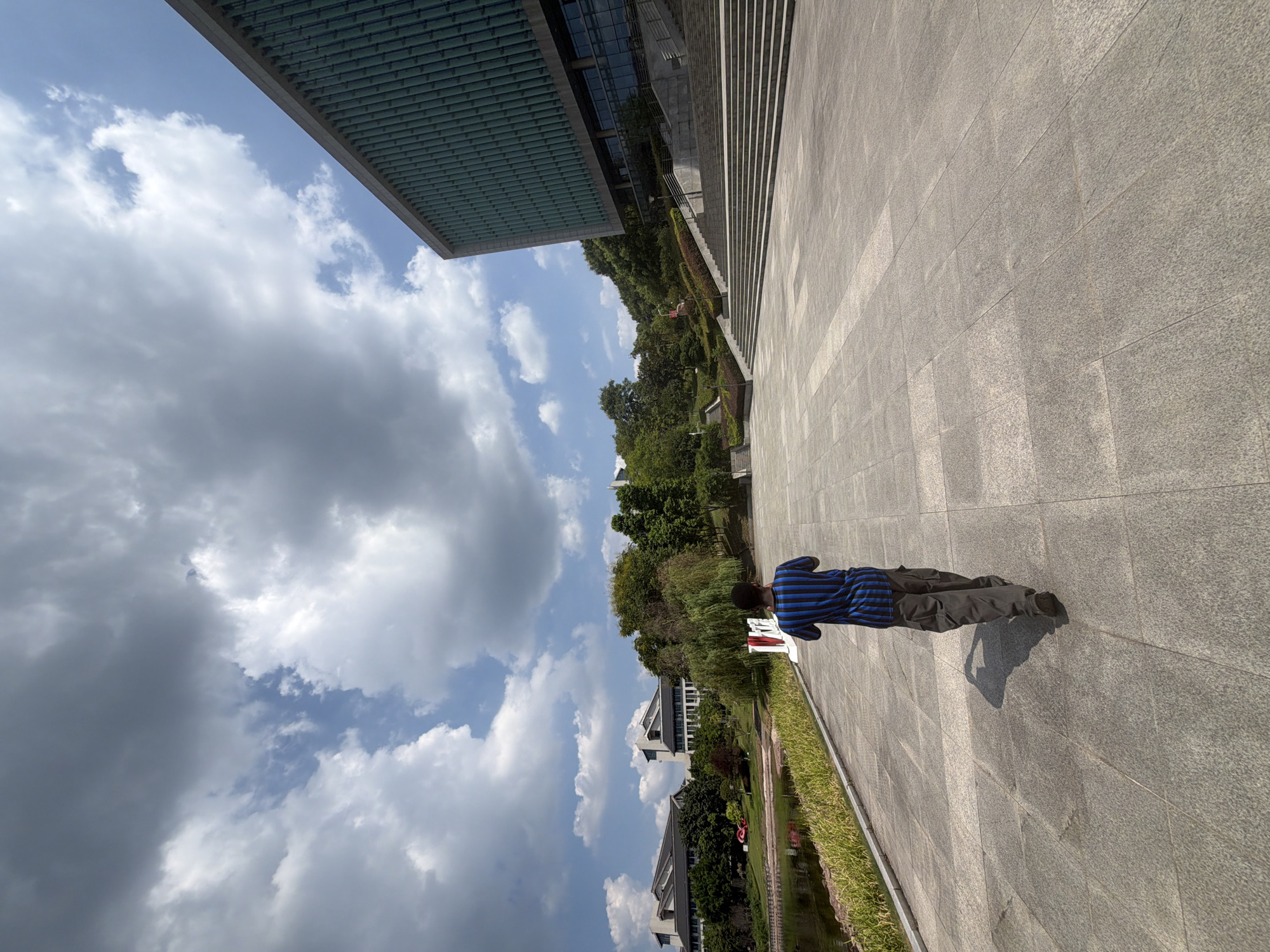 A person walks away from the camera across a large paved plaza. The person is wearing a blue and black striped shirt and grey pants. The plaza is in front of a modern building with a green, ribbed facade and large windows. To the left of the plaza is a body of water with some vegetation along its edge. In the background, there are trees and other buildings. The sky is partly cloudy with a mix of blue sky and white and grey clouds. The scene appears to be on the grounds of a university campus in the Jiangjin District of Chongqing, China.