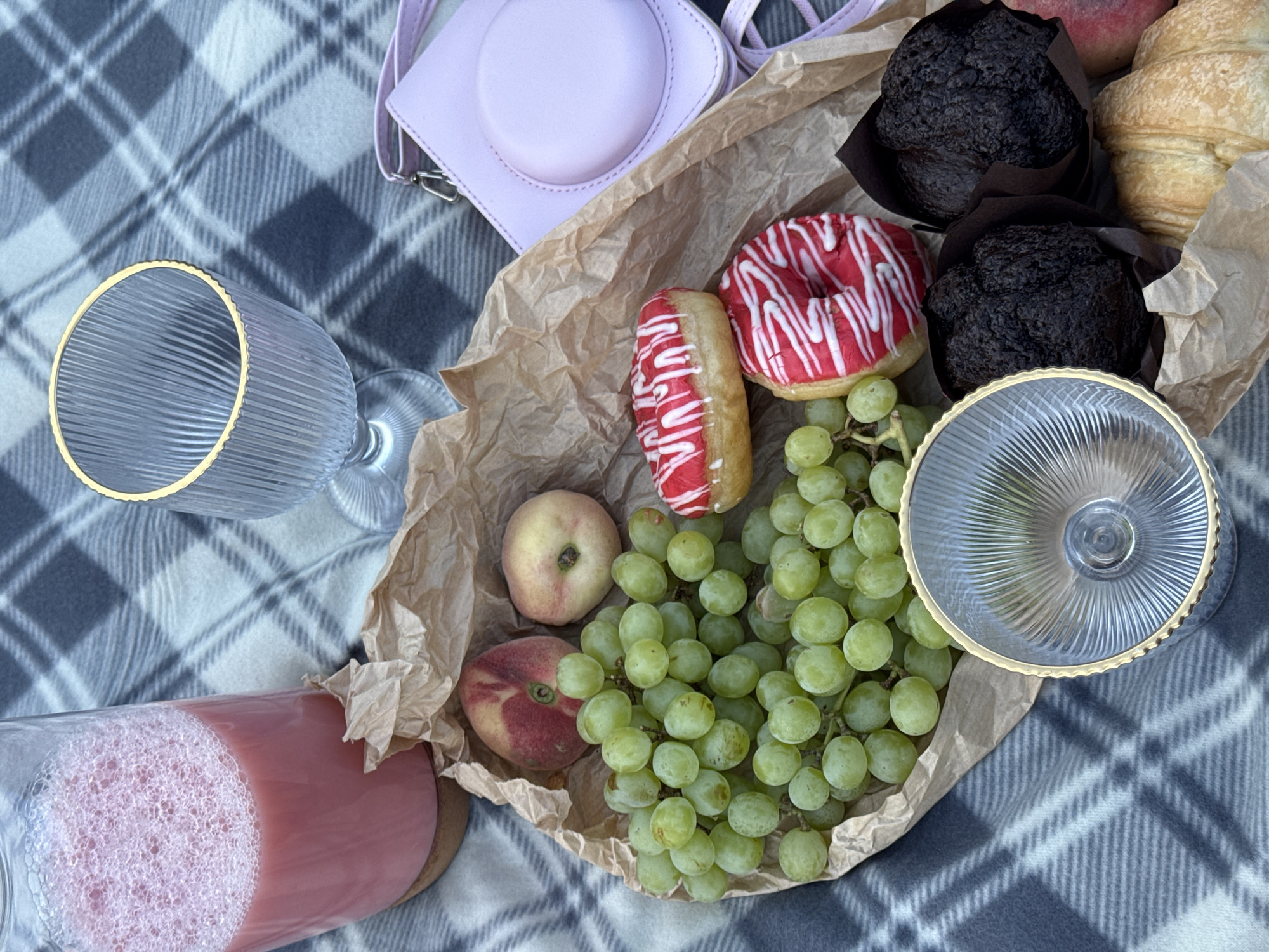 An overhead shot captures a picnic spread on a grey and white checkered blanket. The focal point is a pile of green grapes nestled in brown parchment paper, alongside two red-frosted donuts with white icing drizzle. Two dark chocolate muffins in brown wrappers and two croissants complete the sweet offerings. Adjacent to the treats, a large glass carafe filled with frothy pink liquid sits on a wooden coaster. Two clear, ribbed glasses with gold rims are positioned on the blanket, one empty and upright, the other upside down. A small, pastel pink square camera bag with a strap rests near the glasses. Two peaches, one with red blush and the other lighter, are partially visible among the grapes. The setting is outdoors, implying a picnic scene in Chaikovsky, Russia, though no specific landmarks are identifiable.