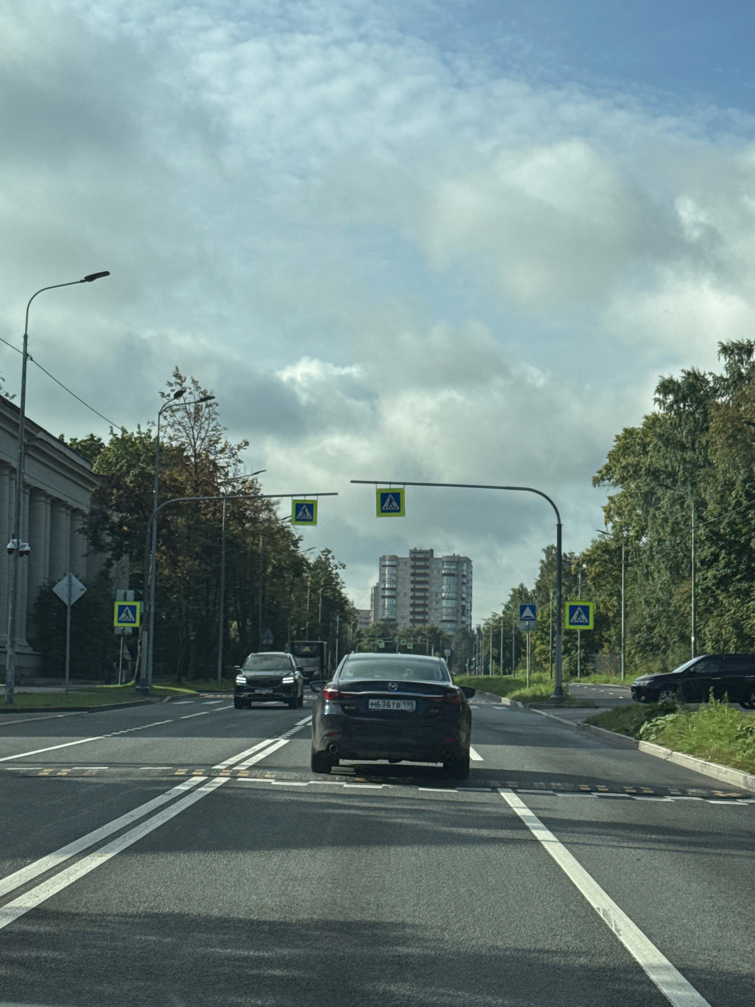 The image shows a view from a vehicle driving on a multi-lane road in a city, likely in Russia given the license plate visible on the rear of the car in the foreground. The road has distinct lane markings and speed bumps. There are several cars on the road, including the one the photo is taken from, a black SUV in front, and other vehicles further ahead. On the left side of the road, there is a large building with columns, and on the right, a dense line of trees. Several pedestrian crossing signs are visible, indicated by yellow square signs with a blue pedestrian symbol, mounted on poles and overhead gantries. In the distance, a tall, modern apartment building is visible. The sky is overcast with scattered clouds, and daylight is present.