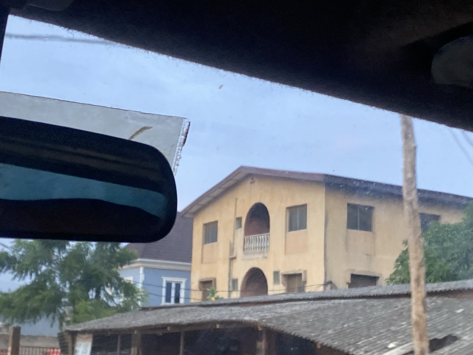 The image, taken from inside a vehicle in Ikorodu, Nigeria, displays a cloudy sky and a view through the windshield. In the foreground, a portion of the car's rearview mirror is visible on the left, with its dark frame and reflective surface. Beyond the mirror, a pale yellow, two-story building with an arched balcony and windows is prominently featured. To the left of this building, a blue, two-story house with white-trimmed windows is partially visible. In front of these buildings, a low-rise structure with a corrugated metal roof is in the mid-ground. Lush green trees are scattered throughout the scene. The overall impression is of a residential or mixed-use area within the city.