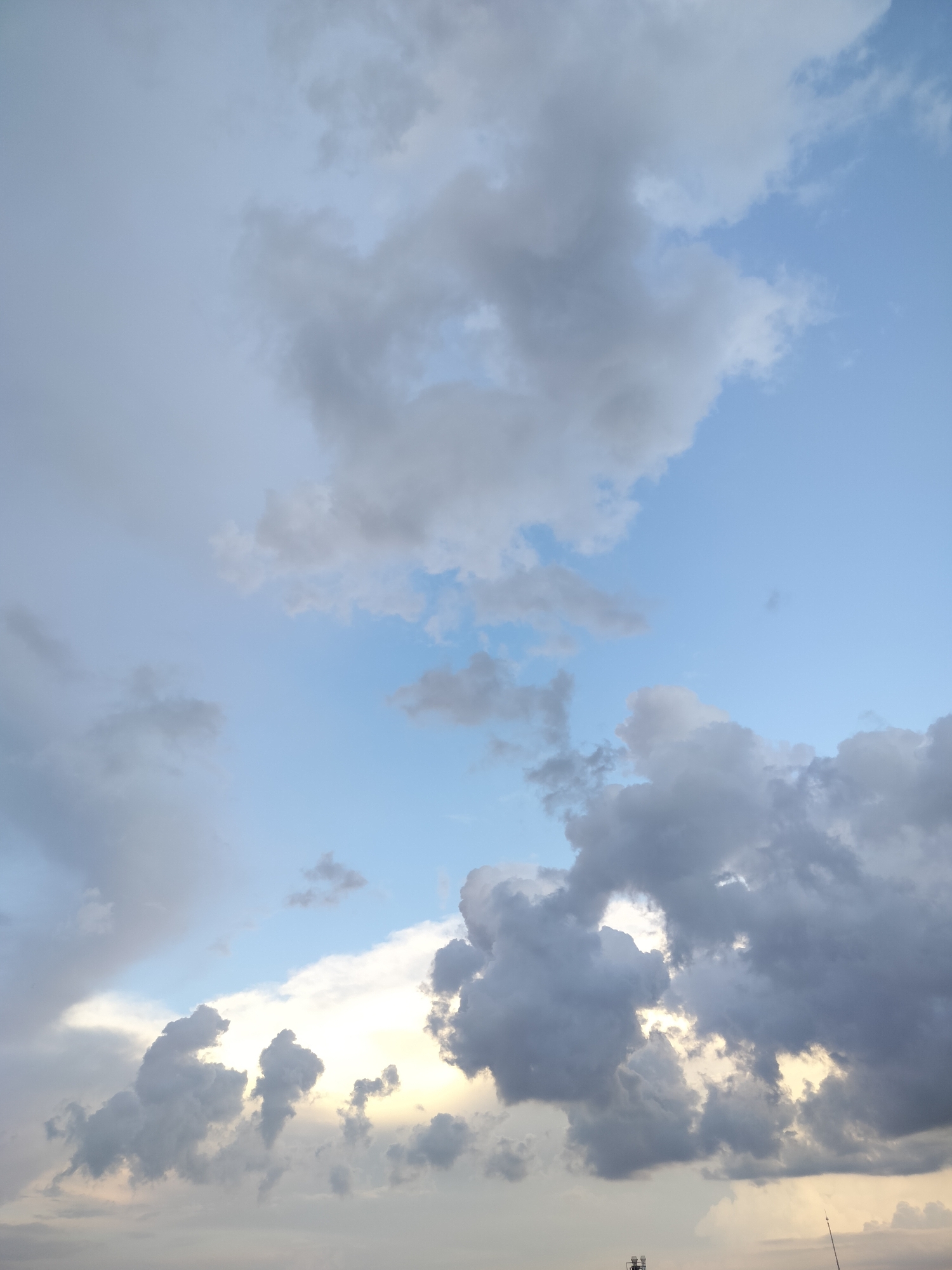 This vertical image displays a vast sky filled with a dynamic cloudscape above Gorno Sahrane, Bulgaria. The upper and middle sections feature a light blue sky with scattered, lighter grey and white clouds. In contrast, the lower and right portions are dominated by larger, denser, dark grey clouds with significant volume. Sunlight, likely from a low-angle sun, breaks through gaps and illuminates the edges of these lower clouds, creating bright, golden-white and yellow hues, particularly towards the bottom-center and lower-right of the frame. This interplay of light and shadow gives the clouds a dramatic and textured appearance.
The time of day appears to be either late afternoon/early evening or early morning, suggested by the warm, directional glow amidst the blue sky and grey clouds. The weather conditions are partly cloudy with significant cloud cover, indicating a changeable sky. No specific human activity or event is depicted. In the very bottom right corner, partially obscured, are a few artificial structures: a slender, tall mast or antenna extending upwards, and a lower structure with visible framework, possibly a communication tower or building edge. No visible text is present in the image.