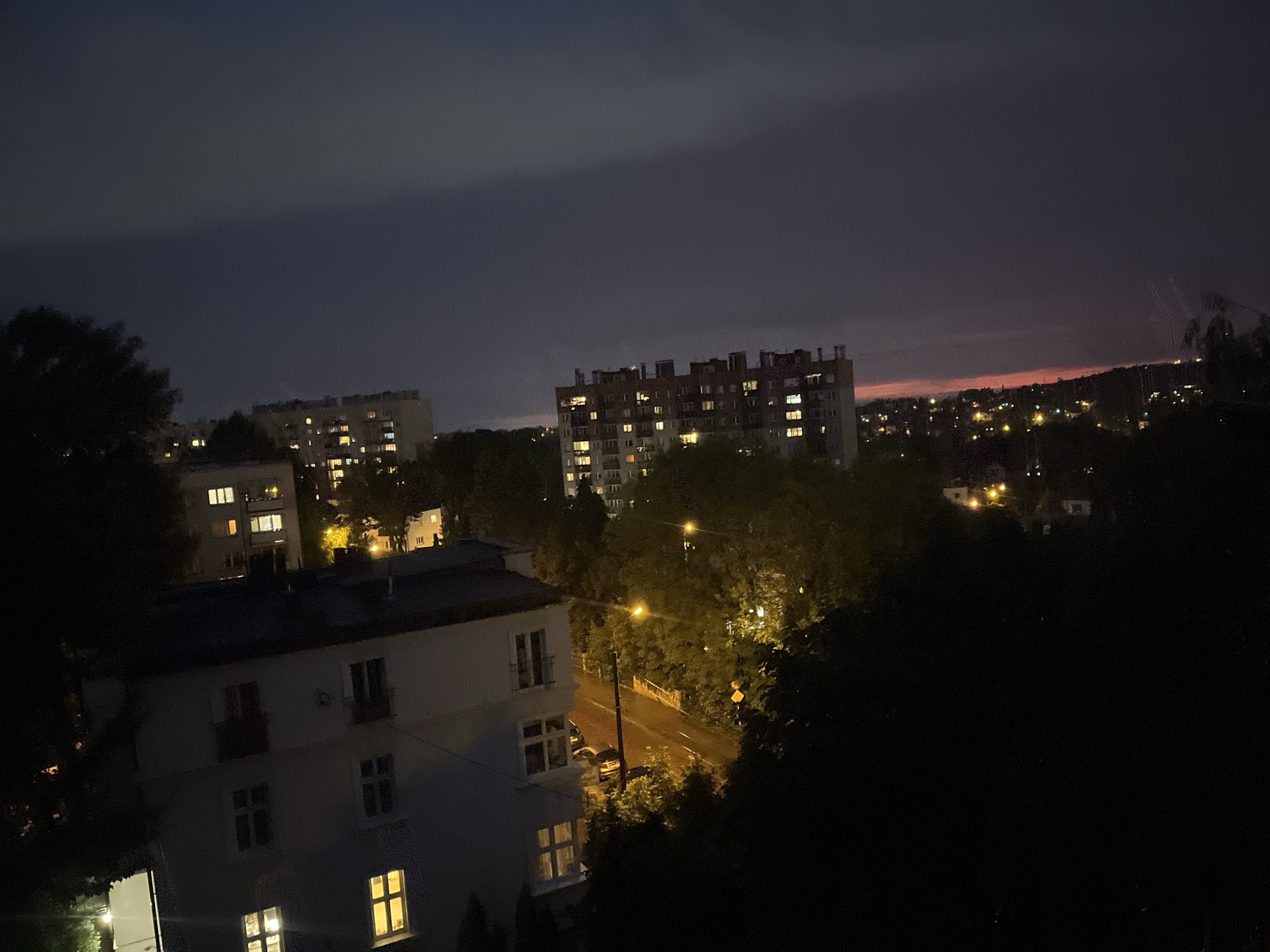 An aerial nighttime view shows a city panorama at dusk. In the foreground, a white building with illuminated windows faces a street with a single streetlamp and parked cars. Further back, other residential buildings are visible, some with lights on. The sky is dark, transitioning to a faint reddish hue near the horizon, indicating the last light of sunset. Trees are scattered throughout the scene, with some silhouetted against the sky and others illuminated by distant lights. The overall impression is a quiet urban evening.