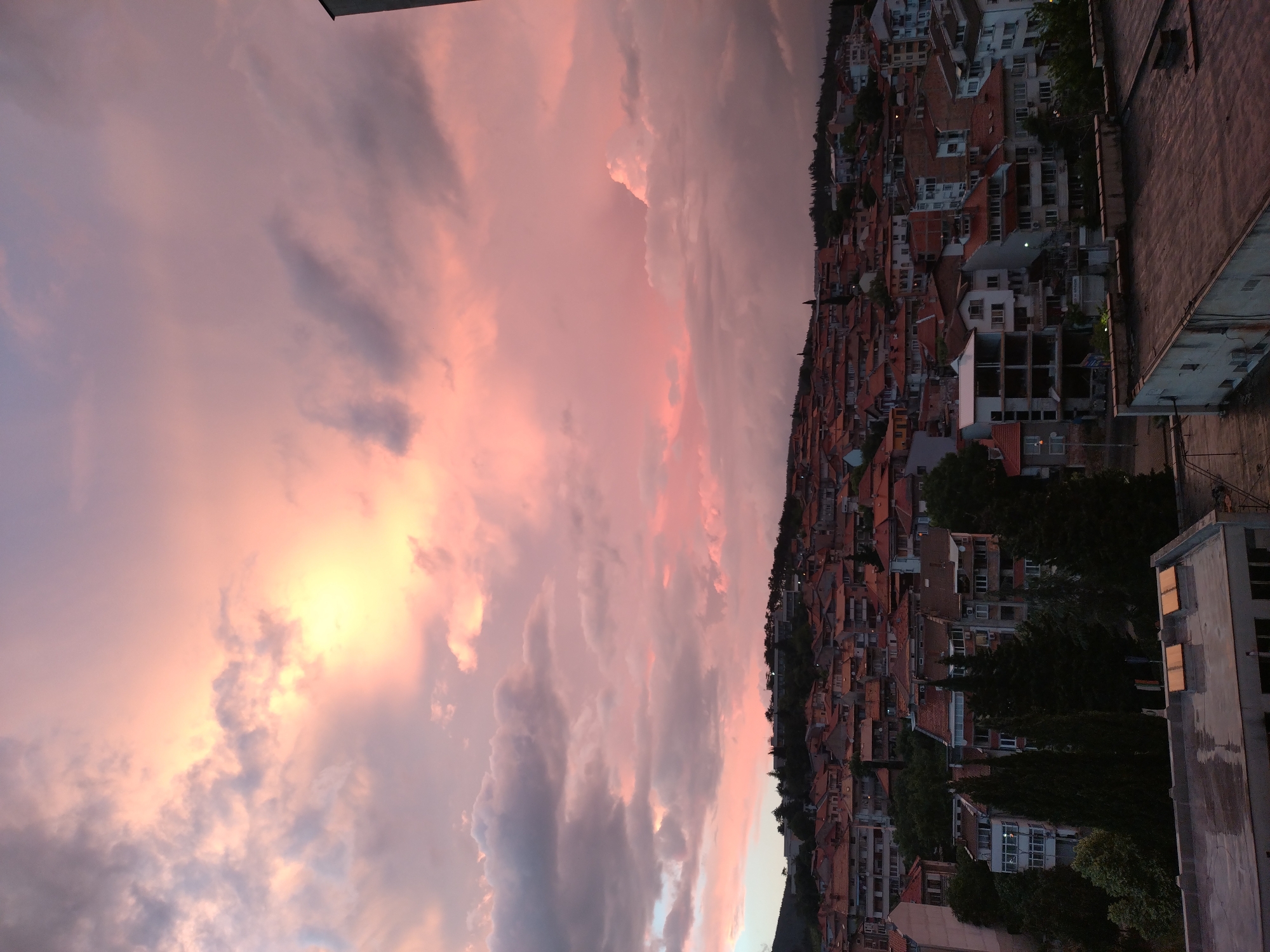 The image shows a view of the city of Stara Zagora, Bulgaria, under a dramatic, colorful sunset sky. The sky is filled with pink, orange, and gray clouds, with a bright area indicating the setting sun. Below the sky, a dense urban landscape stretches across the frame. This landscape is predominantly composed of buildings with red-tiled roofs. The buildings vary in size and appearance, creating a complex pattern across the urban scene. Some buildings are taller and more modern, while others appear older and more traditional. Dark green trees are scattered among the buildings, providing a contrast to the red roofs and adding depth to the image. In the foreground of the image, there is a rooftop with some taller buildings near the viewer.