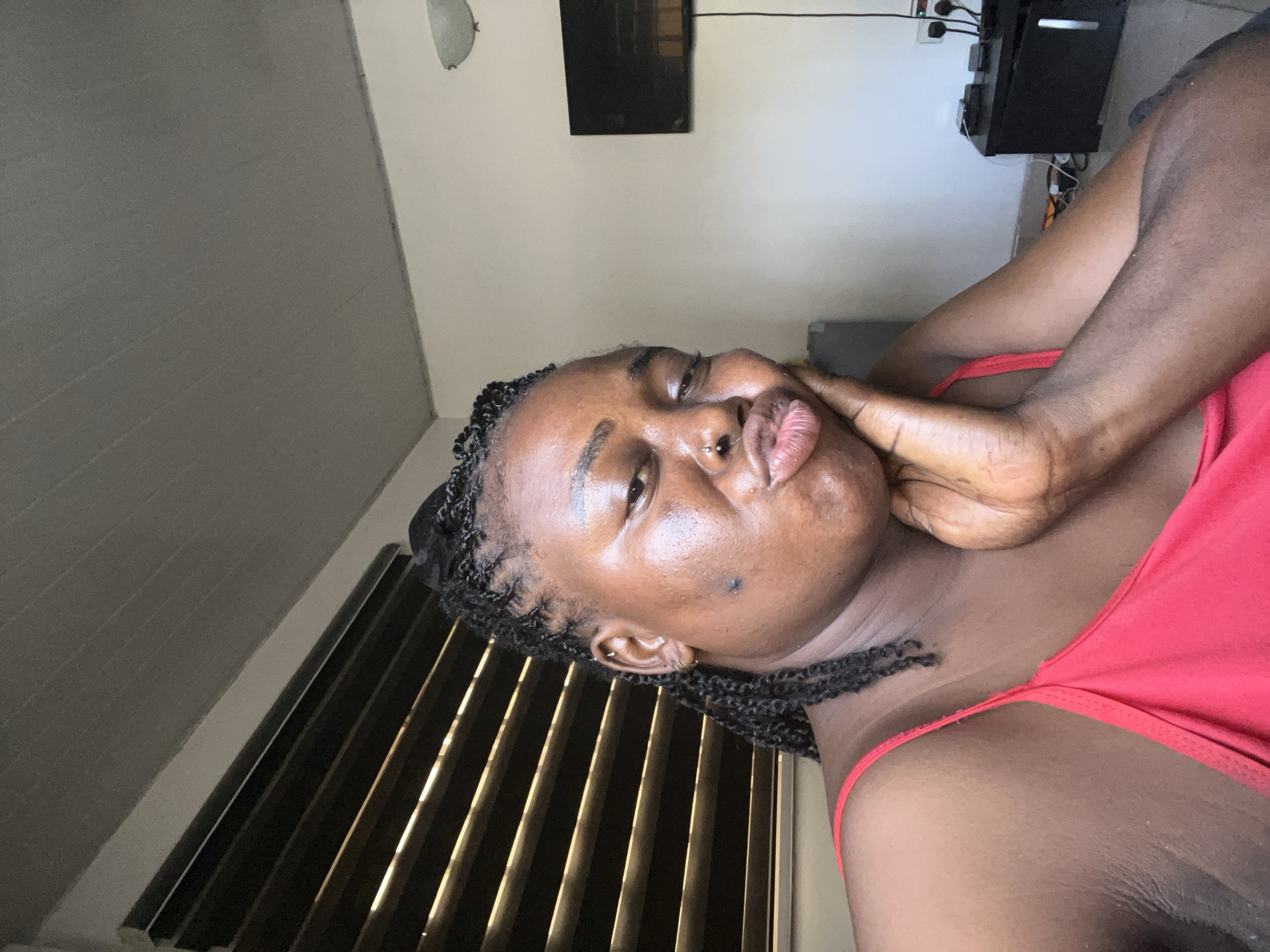 A close-up, angled shot captures a young Black woman with braided hair and a nose ring. She is wearing a red tank top and appears to be indoors. Her left hand is touching her cheek, and she has a playful expression with her lips puckered. The background suggests a domestic setting with a wall, a window with blinds, and some electronic devices mounted on the wall. A framed picture or television is visible on the wall. The lighting indicates it is likely daytime. The overall mood is casual and intimate. There is no visible text in the image.