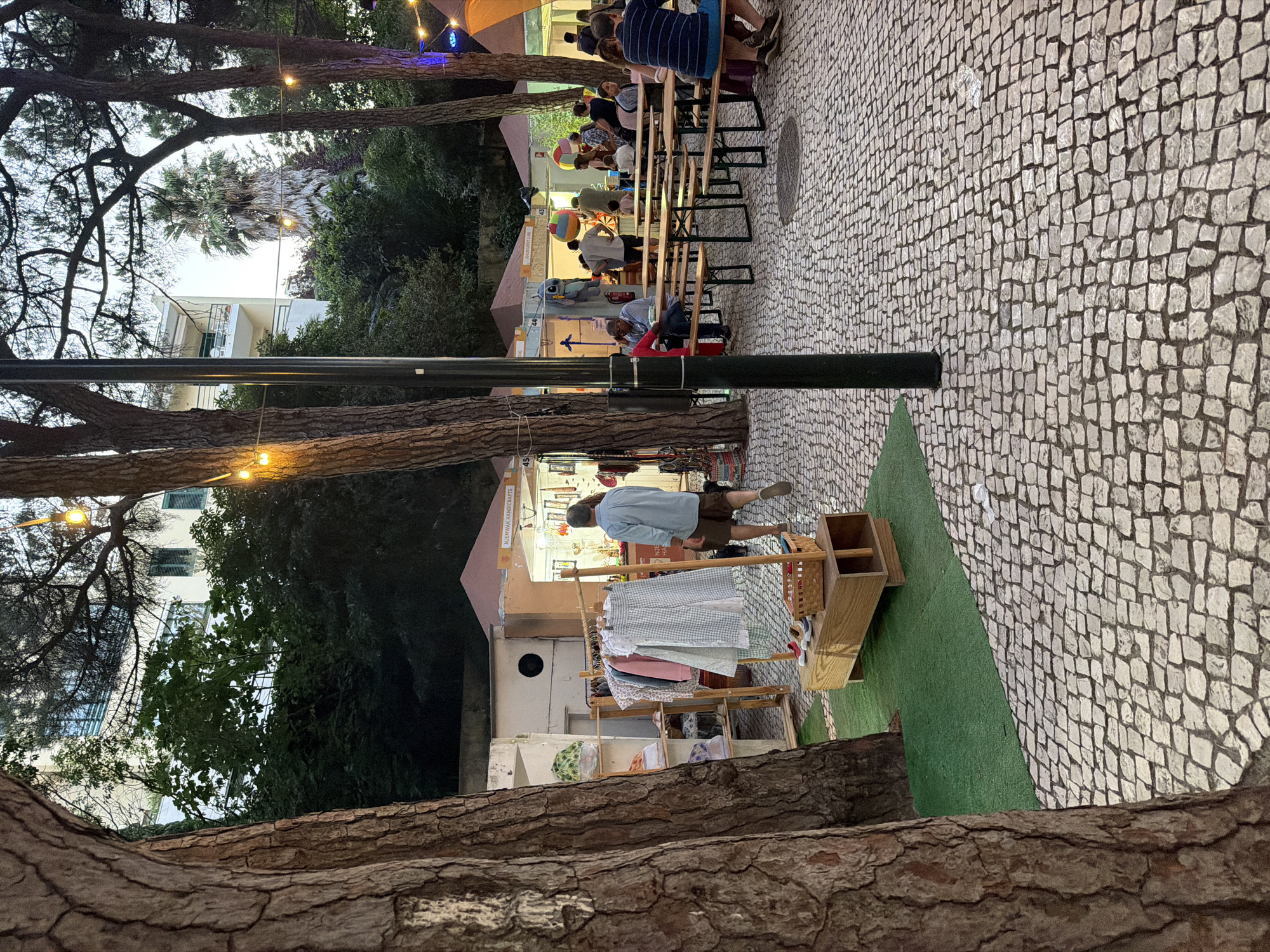 This is a medium-shot, eye-level photograph taken at dusk in Cascais, Portugal, at a street market with a cobblestone ground and trees. In the foreground, the rough bark of a large tree trunk is on the left side, obscuring part of the scene. To the right of the tree, a wooden clothing rack displays several garments, including a light blue and white checkered dress, a pink shirt, and a white and blue patterned shirt. A wooden bench with a basket containing shoes sits at the base of the rack.
Beyond the clothing rack, several market stalls are set up, some with signage like "NJEMWAK HANDICRAFTS." People are milling about and sitting at long wooden picnic tables. String lights are visible overhead, adding to the evening ambiance. In the background, a multi-story building with balconies and windows is partially visible through the trees, along with more greenery and a tall palm tree. The overall scene depicts a lively outdoor market environment in the city.