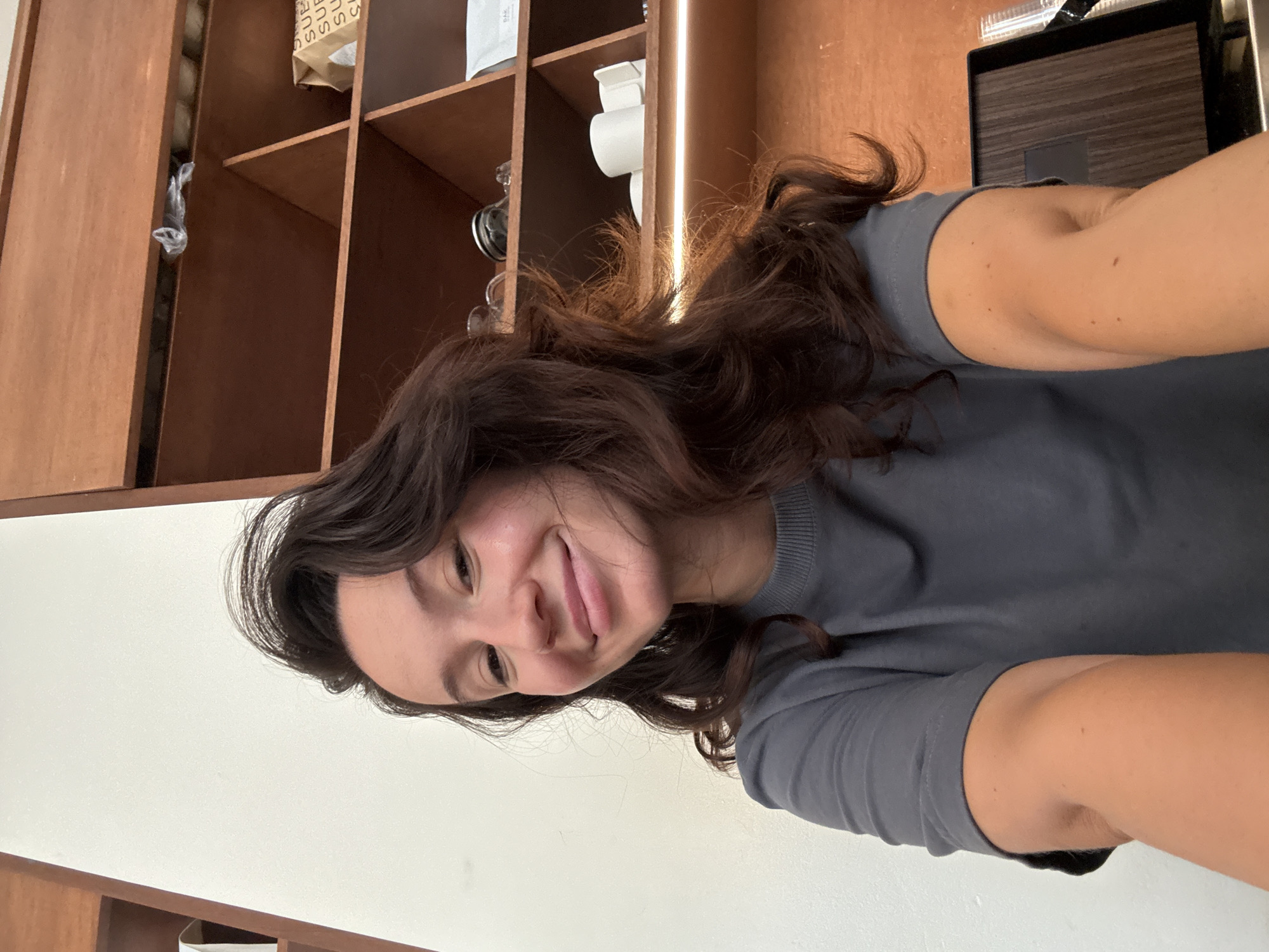 A young woman with long, dark, wavy hair smiles into the camera. She is wearing a dark grey t-shirt. She is positioned in front of a wooden shelf unit with various items on it, including cups, a bag, and what appears to be a plastic bag. The background on the left is a plain white wall. The image appears to be taken in an indoor setting.