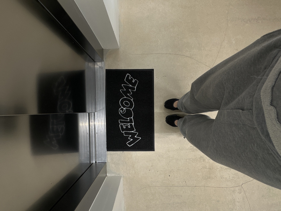 The image presents a first-person, downward-facing view of a person's lower body standing in an interior space within the city of Paris, France. The individual wears grey sweatpants and black footwear with a distinct split-toe design. Directly in front of the person's feet, a rectangular black doormat features the word "WELCOME" in white, stylized, rounded lettering. Beyond the doormat, a set of closed metallic, reflective doors, consistent with an elevator or modern building entrance, occupies the upper portion of the frame. Horizontal metallic tracks are visible at the base of these doors. The reflective surface of the doors displays a blurred, inverted reflection of the "WELCOME" text. The floor, extending from the person to the metallic doors, is a light-toned surface, possibly concrete or large tiles, exhibiting minor surface cracks. White wall sections frame the metallic entrance on either side.
