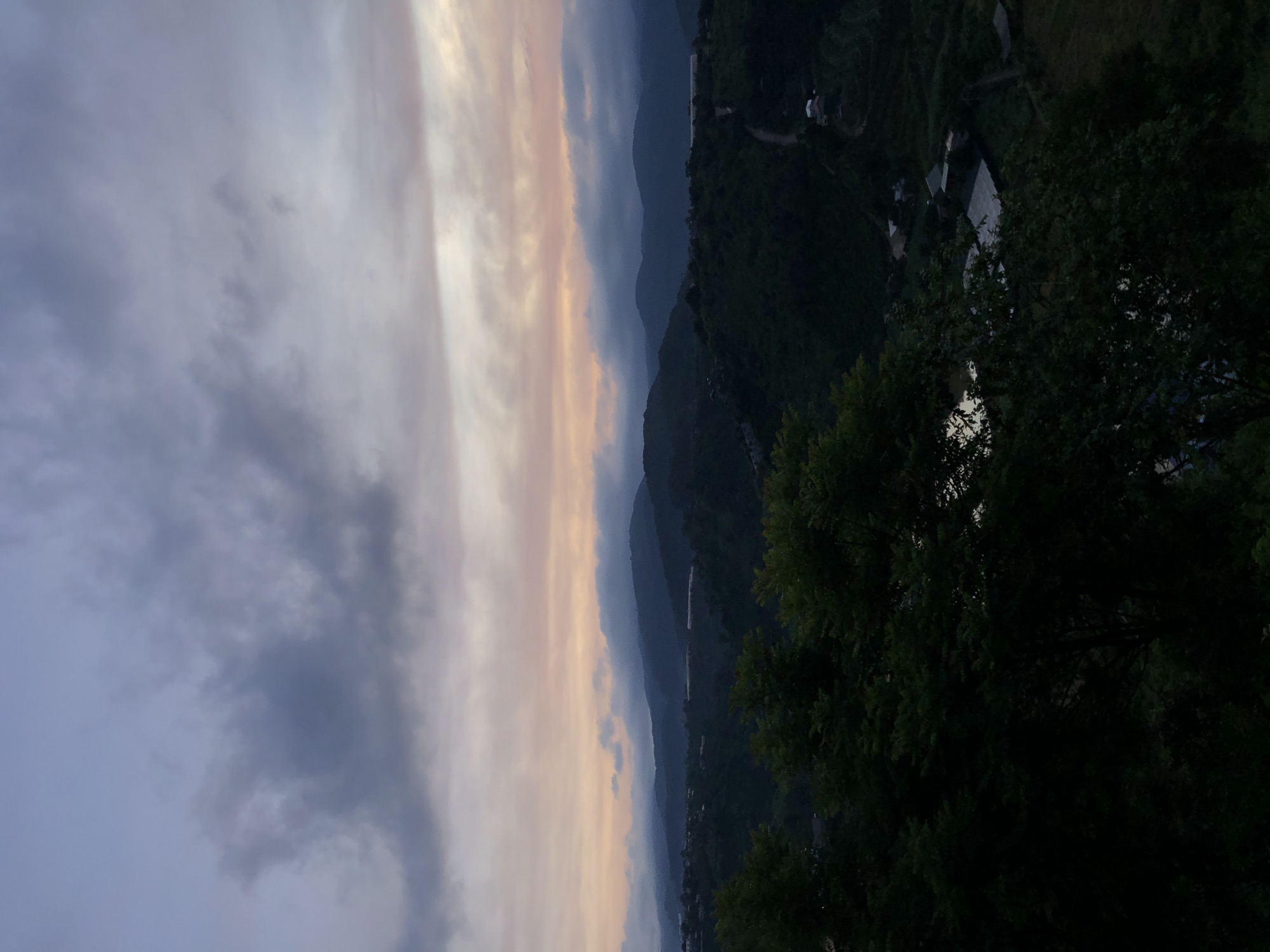 The image captures a panoramic view of a rural landscape, likely in or near Thôn 3, Vietnam, during twilight. The sky is filled with dramatic, dark clouds that transition into softer hues of pink and orange near the horizon, suggesting either sunrise or sunset. Below the sky, a series of rolling hills covered in dense, dark green vegetation dominate the midground and background. The topography appears mountainous, with layered peaks fading into the distance.
Scattered across the hillsides, especially in the midground and lower right, are signs of human settlement. Several small buildings with light-colored roofs are visible, some appearing to be houses or agricultural structures. Narrow, winding paths or roads snake through the terrain, connecting these scattered dwellings. In the foreground, the dark silhouettes of leafy trees frame the scene, obscuring portions of the landscape and adding depth. The overall impression is one of a serene, rural evening in a hilly region, with the remnants of daylight illuminating the sky above a shadowed landscape.