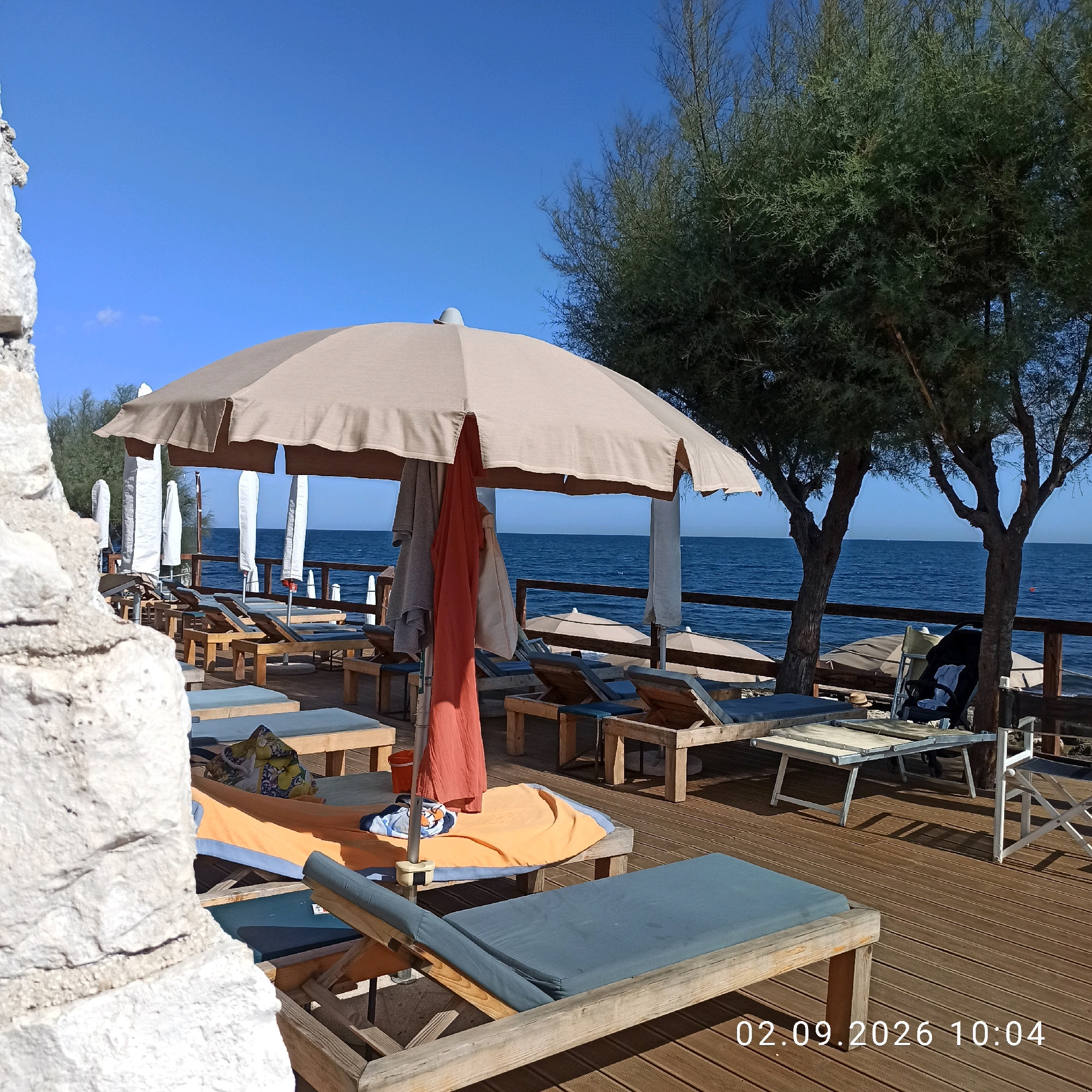 The image depicts a beachside setting in Molfetta, Italy, with a focus on a wooden deck area furnished with lounge chairs and umbrellas. The scene is oriented towards the sea, which is calm and blue under a clear sky.
In the foreground, a large, light brown umbrella is prominent, with draped fabrics hanging from its pole, including a coral-colored towel and a grey garment. Several wooden lounge chairs with blue cushions are arranged on the deck. To the left, a large, textured white rock formation partially obscures the view.
The wooden deck extends into the background, where more lounge chairs and umbrellas are visible. A wooden railing separates the deck from the sea. Trees with dense green foliage are present, one on the right side of the image, contributing to the natural environment. The sea is a deep blue, with a few distant white caps suggesting slight waves.
In the middle ground, a black stroller is positioned near some lounge chairs. A collapsible table with a metal frame and a light-colored top is also present on the deck. The overall impression is one of a well-maintained beach resort or club area, prepared for relaxation.
The bottom right corner of the image displays a timestamp: "02.09.2026 10:04".
