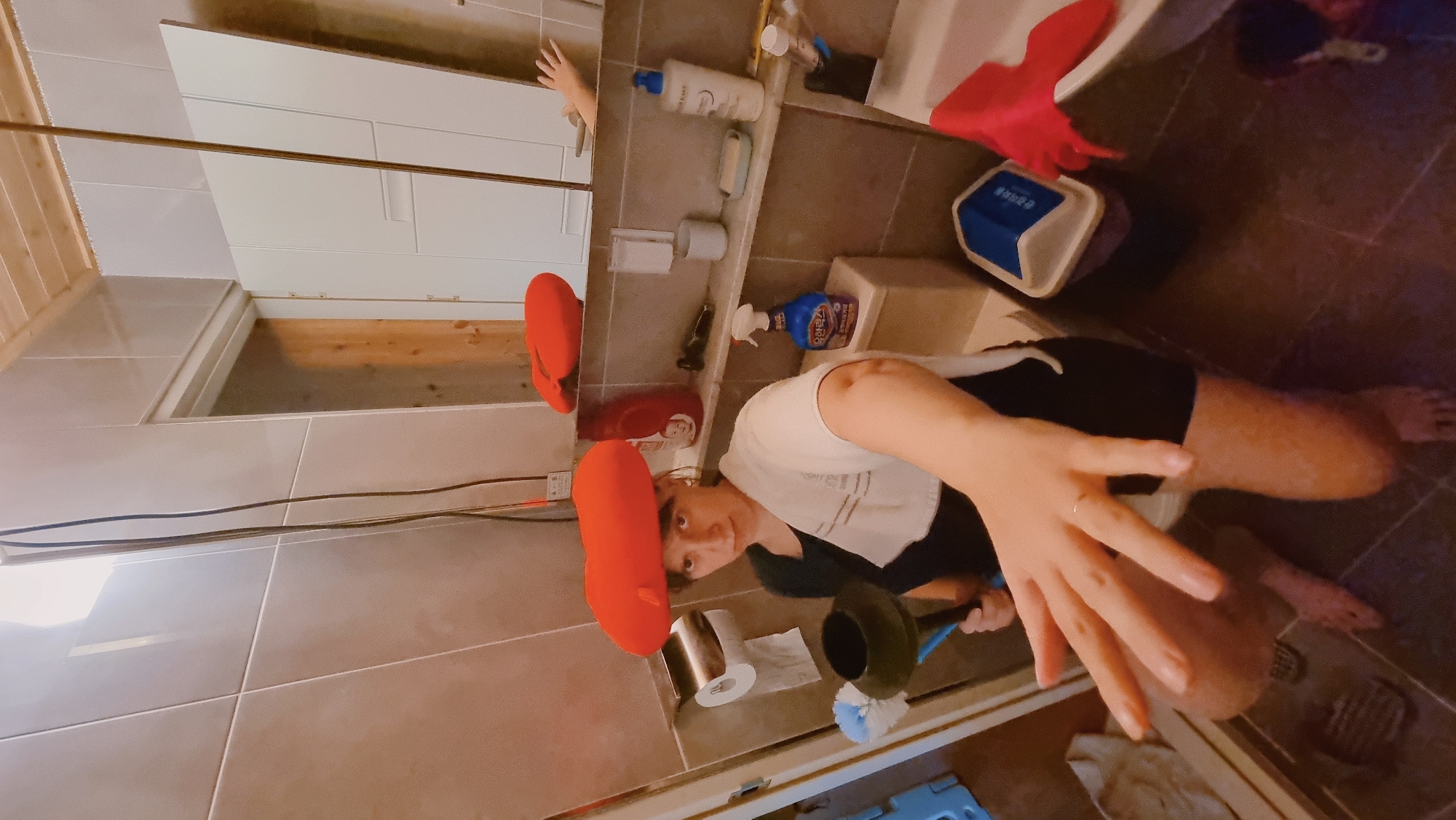 In a bathroom located in Dunnae-myeon, South Korea, a person wearing a white towel and black shorts is cleaning a toilet with a toilet brush and plunger. A red toilet brush holder sits next to the toilet. The bathroom features a mirror, tiled walls, a white bathtub, and various cleaning supplies on a shelf. The floor is tiled.
