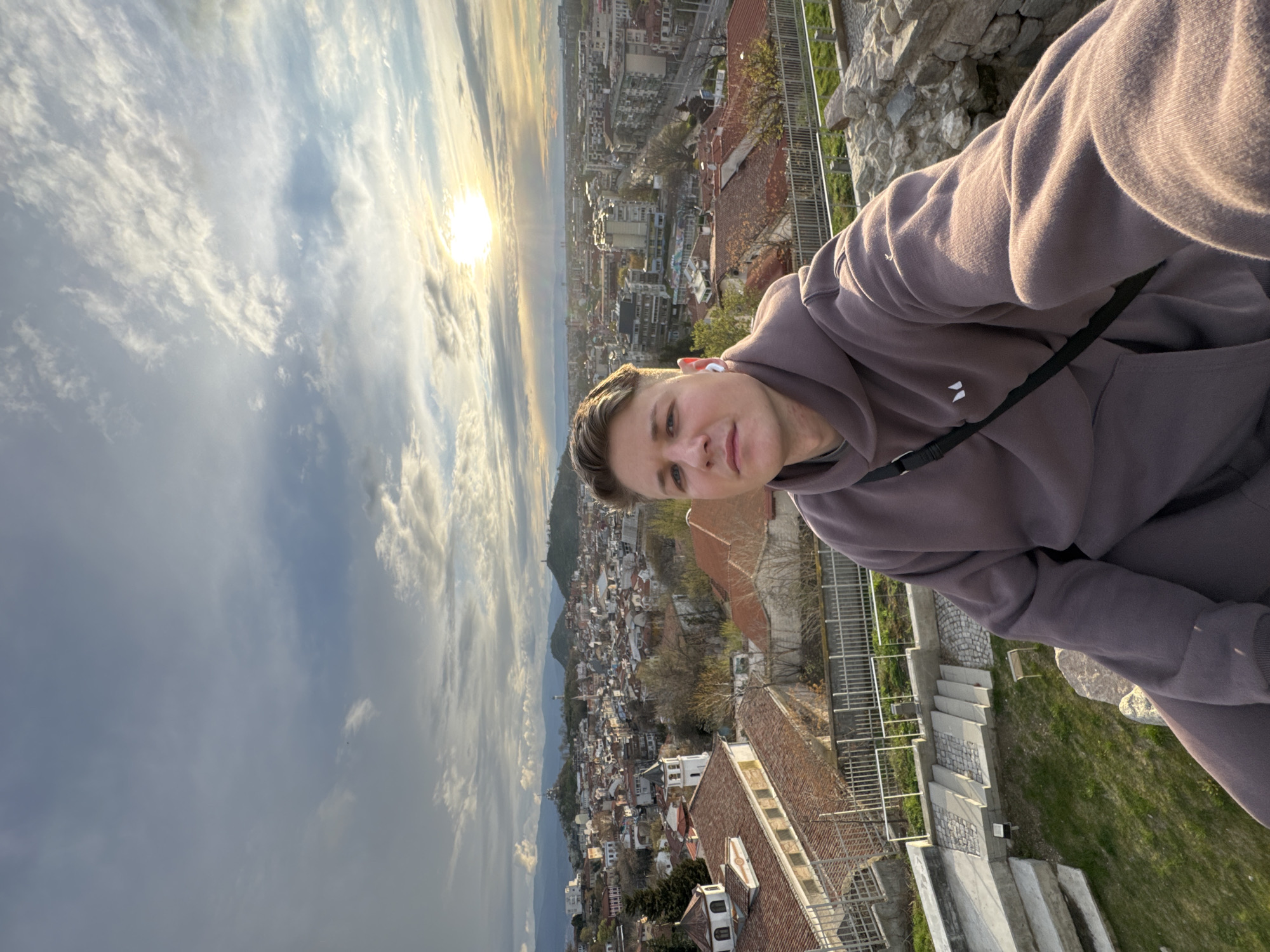 A vertically oriented selfie shows a young man standing on an elevated stone structure overlooking the city of Plovdiv, Bulgaria, at sunset. The man wears a brown hoodie and white wireless earbuds. A black strap of some kind crosses his torso. In the background, the cityscape stretches out, with various buildings and rooftops visible, including likely a few from the Old Town. The sky is filled with clouds and the setting sun. The picture is taken from Nebet Tepe.