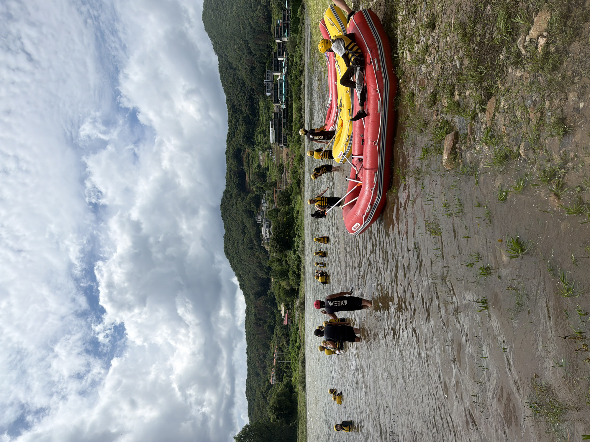 A group of people, most wearing yellow life vests and some with helmets, are in a shallow river, some standing and others wading. Two red inflatable rafts are positioned in the foreground, one partially in the water and one on the rocky bank. A yellow inflatable raft is further out in the water. In the background, a lush, tree-covered hillside rises, with buildings scattered on its slopes and a road visible. The sky is overcast with white and gray clouds. This scene is located in Seo-myeon, South Korea.