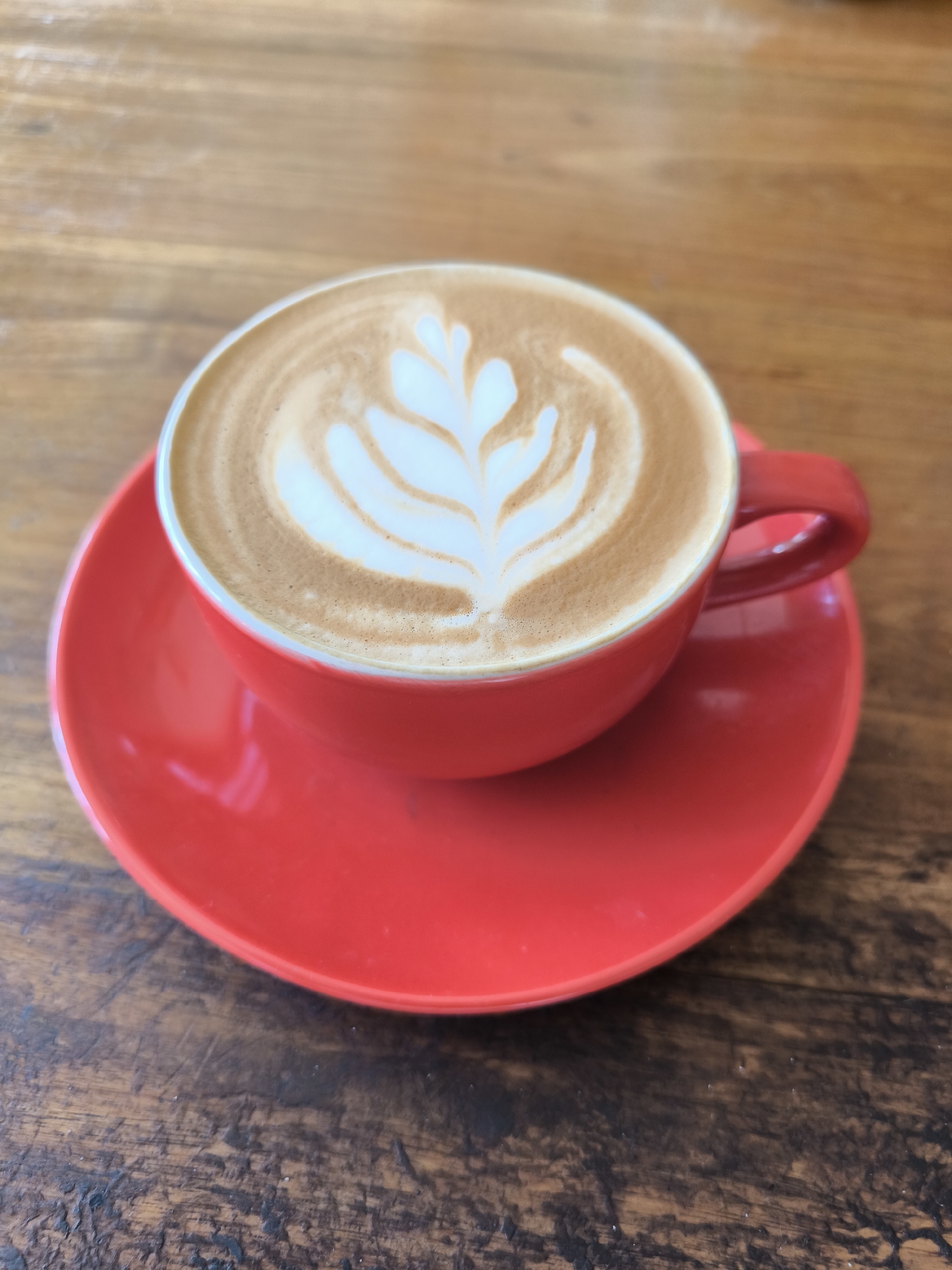 A red cup of latte with latte art sits on a matching red saucer atop a wooden table. The setting is Unknown, Indonesia.