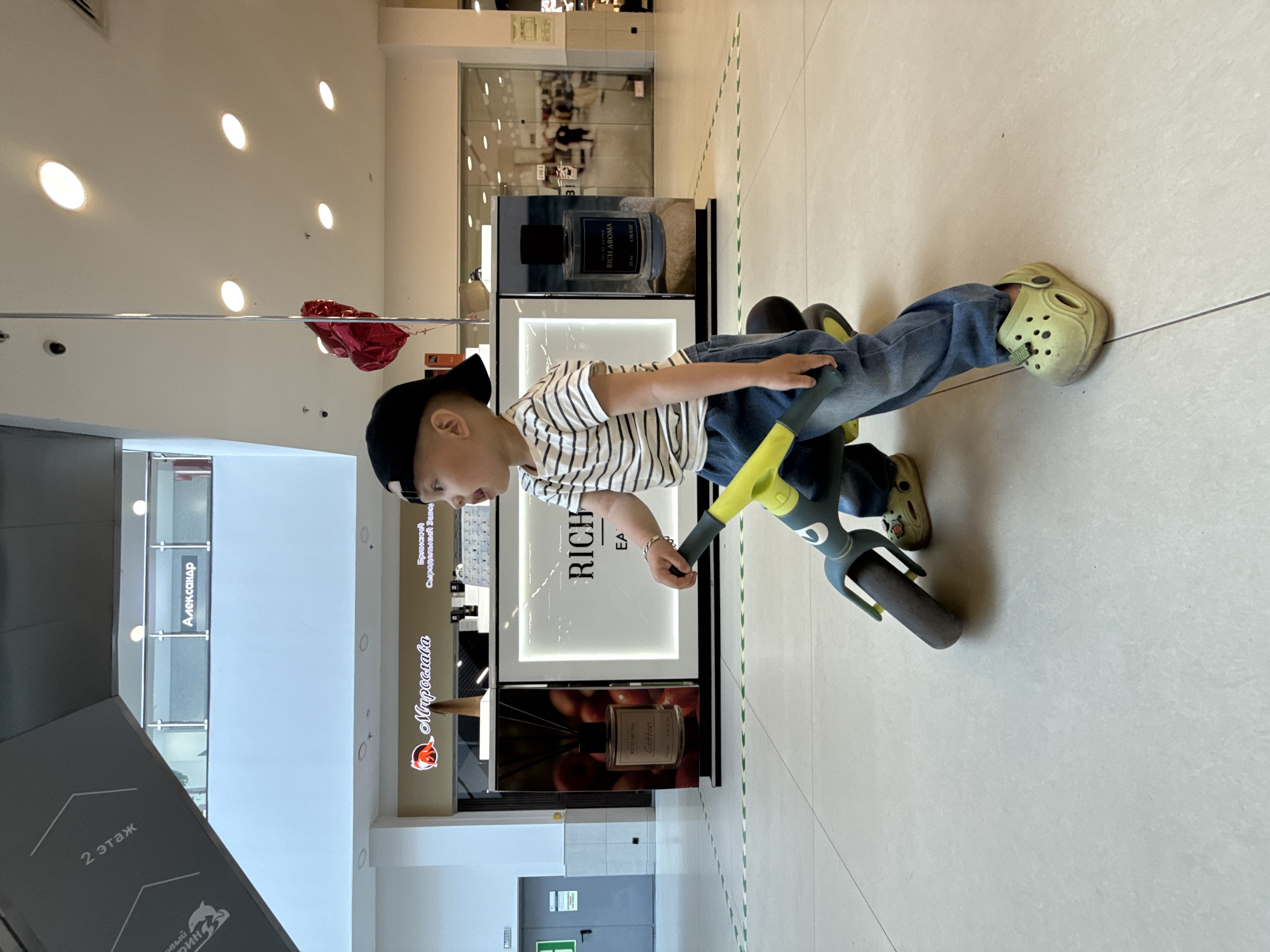 A boy wearing a black cap, a striped shirt, and jeans, with green shoes, is riding a blue and yellow tricycle in what appears to be a shopping mall in Rusanovo, Russia. There is a sign on the left that says "Мирослава" (Miroslava) and "2 этаж" (2nd floor). Adjacent to the boy is a promotional display for "RICH AROMA" perfume. Behind the display is a store front with the name "Александр" (Alexander) visible. The mall has white tile floors and bright overhead lighting.