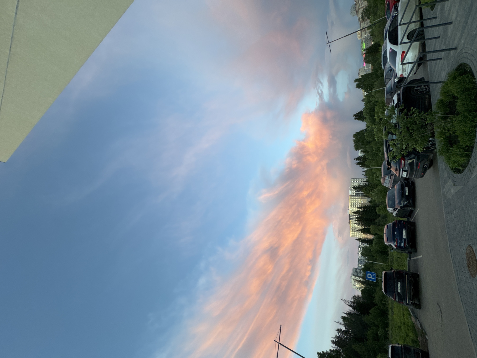 The image captures a view from what appears to be an urban area, likely a parking lot adjacent to a building. The focus is on the sky, displaying a dramatic sunset with elongated, peach-colored clouds stretching across a blue sky. On the right side of the frame, a row of parked cars lines the edge of the image, suggesting a parking area. A line of trees and glimpses of city buildings are visible behind the parked cars, indicating an urban environment. Pavement and a sidewalk with some patterns are noticeable on the right side of the image. The image is rotated counterclockwise. While the image doesn't provide concrete clues, the metadata mentions that the image was taken in Izhevsk, Russia.