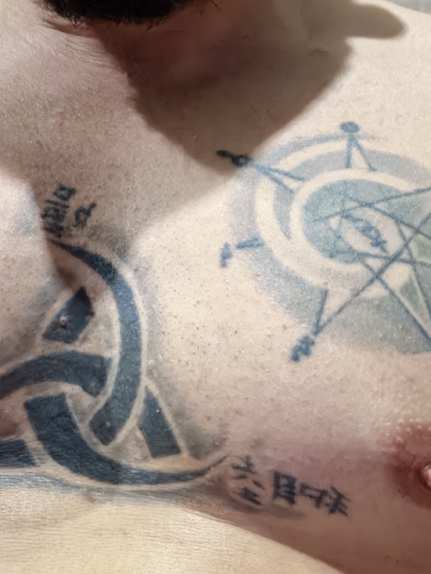 This image shows a close-up view of a person's torso. The skin is light-colored with some visible pores and imperfections. There are several tattoos visible on the chest. One tattoo is a Celtic knot design, another is a circular design resembling a compass or alchemical symbol, and there appear to be two sets of characters or symbols, potentially in a foreign language. A nipple is visible on the right side of the frame. The image is well-lit, but the angle is close and intimate.
