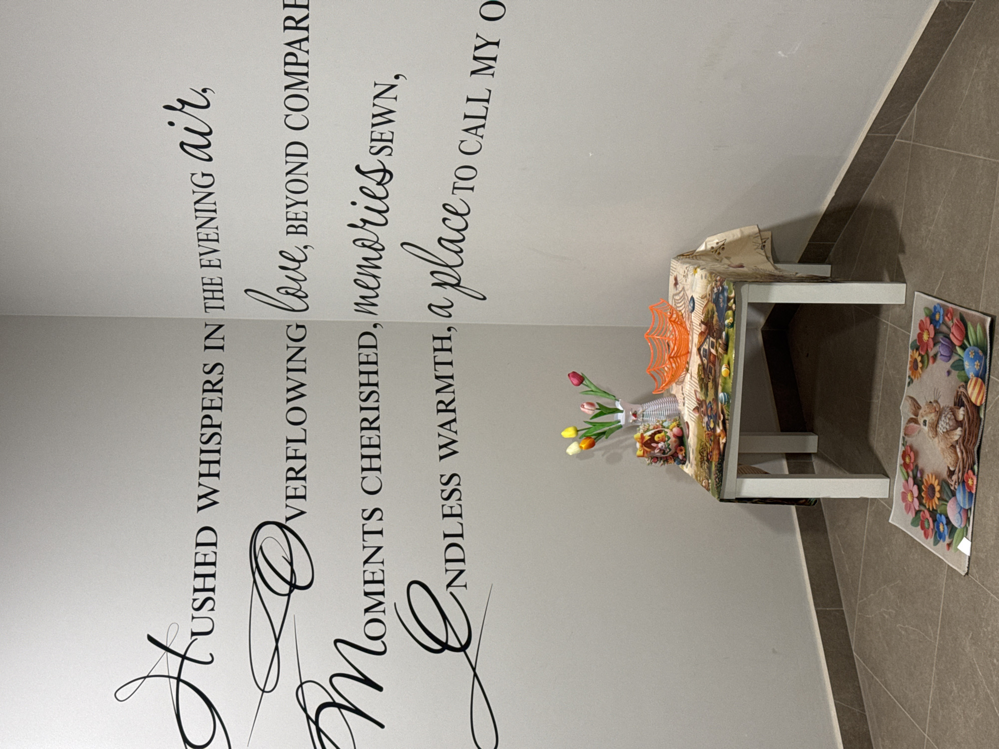 A room has a white wall with black text on it. In the corner is a small white table with a patterned tablecloth. On the table are flowers and an orange decoration. A patterned mat is on the floor.