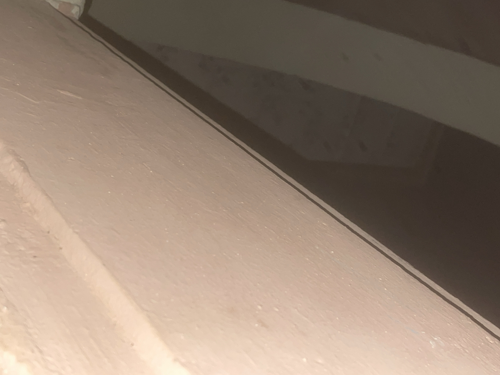 The image shows a close-up, low-angle view of a wall with a textured, light pinkish-beige surface. A thin, dark line runs diagonally across the image, marking the edge of the wall or a separation from another surface. The wall appears to be an interior surface, possibly a corner or an architectural detail. The lighting is dim and casts shadows, suggesting an indoor environment with limited light sources. The texture of the wall is rough and slightly uneven, with small specks or imperfections visible. The lower portion of the image is darker and shows a shadow or a different, darker surface that is out of focus. No people, specific objects, or activities are discernible. The scene is devoid of any specific identifying features that would allow for pinpointing its location in Howayi, Nigeria, or any other city.