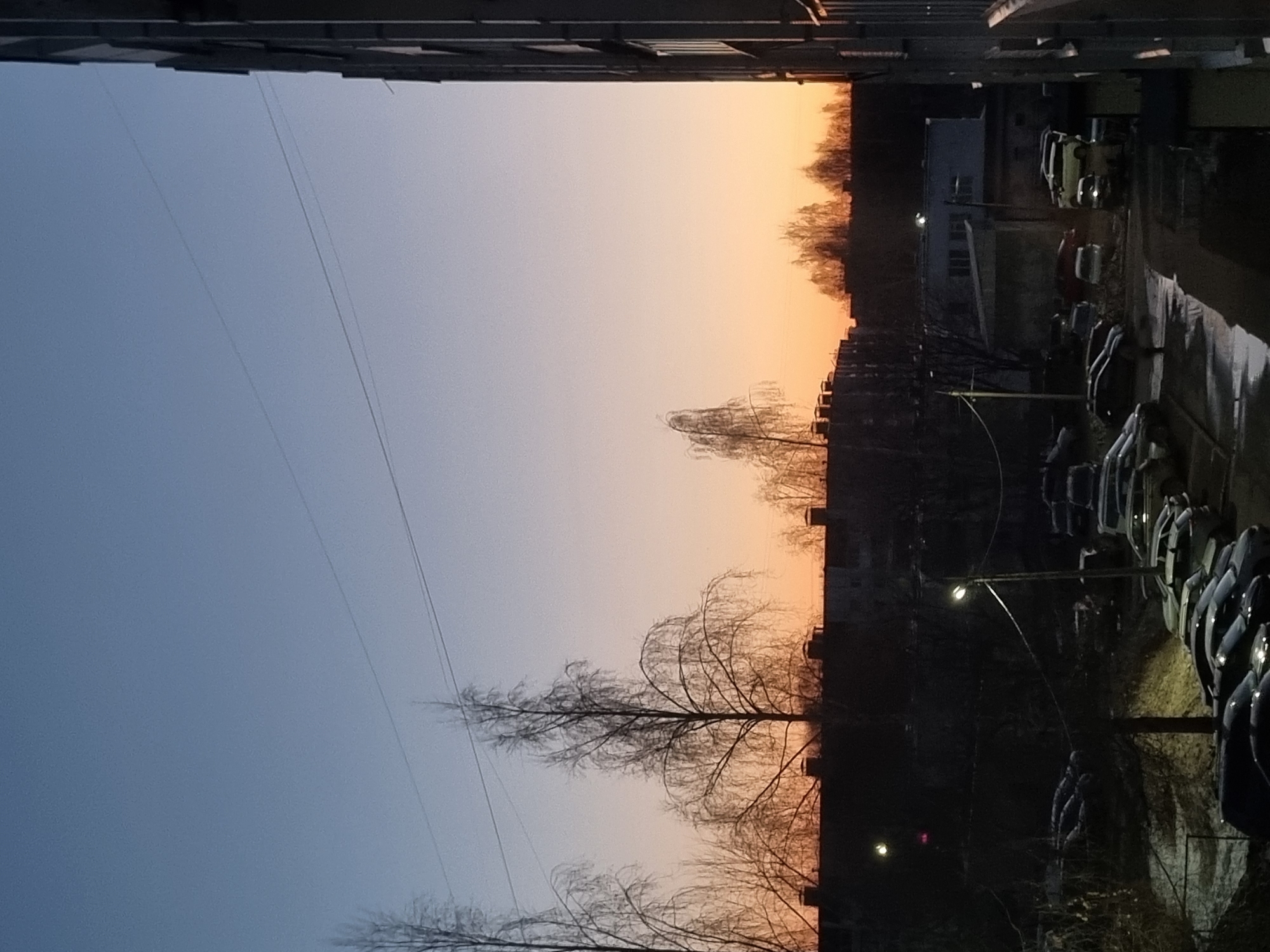 The image captures a twilight view in Chaikovsky, Russia, framed by the edges of apartment buildings. The sky exhibits a gradient, transitioning from a soft blue to a warmer orange hue near the horizon, suggesting either sunrise or sunset. Silhouetted trees with bare branches stand against the colorful sky. A row of parked cars lines the lower right portion of the image, illuminated by streetlights. Adjacent to the cars are buildings, which also appear silhouetted due to the lighting conditions. Electrical wires run diagonally across the sky on the left.