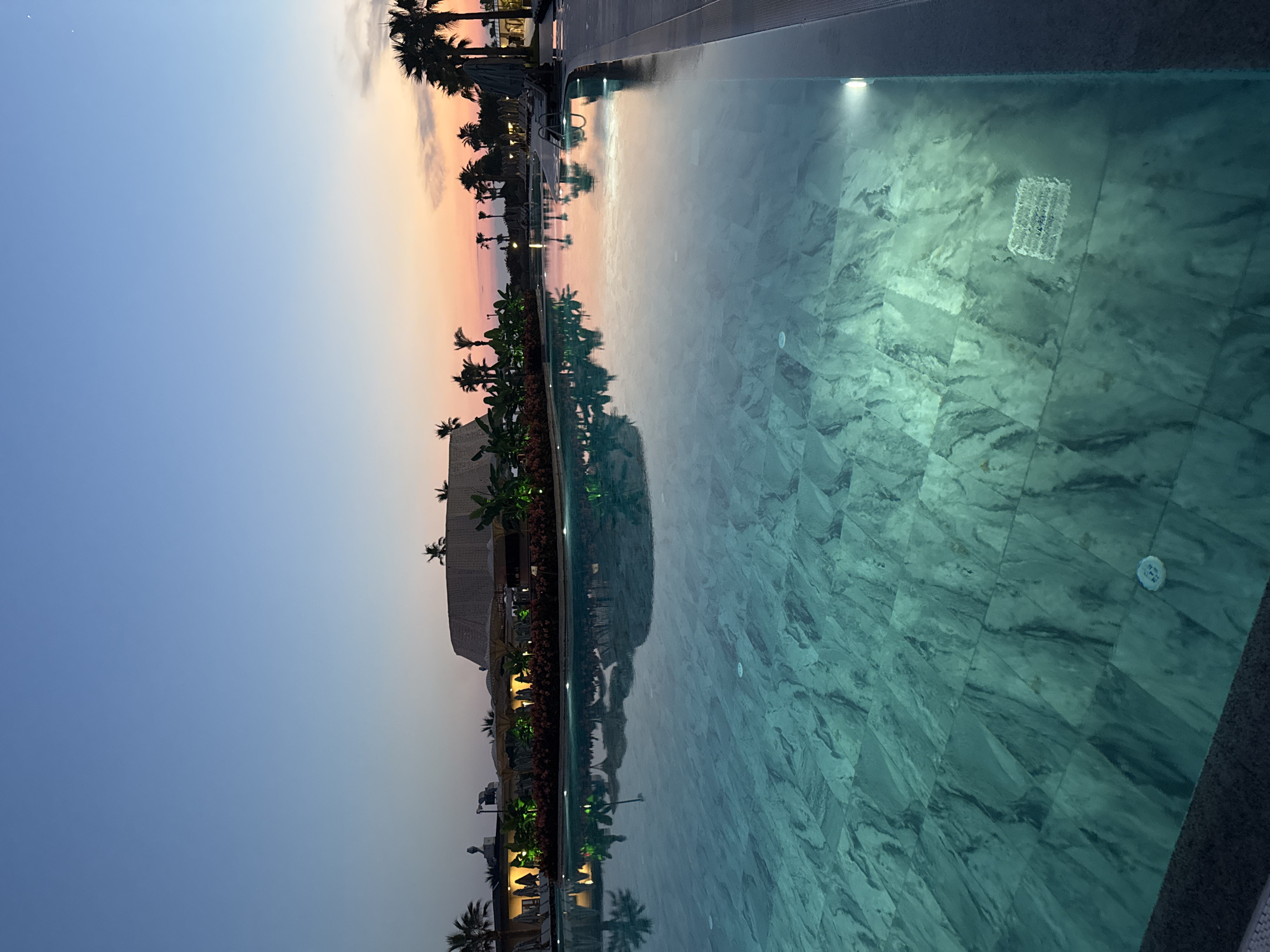 The image shows a serene swimming pool at dusk, with the sky transitioning from a clear blue to hues of pink and orange. Palm trees and resort buildings with ambient lighting are visible in the background, reflecting on the water's surface. The pool itself is illuminated from within, highlighting the marbled tiles of its floor. The scene evokes a sense of tranquility and relaxation, typical of a resort setting in Manavgat, Turkey. There are no people visible in the image.