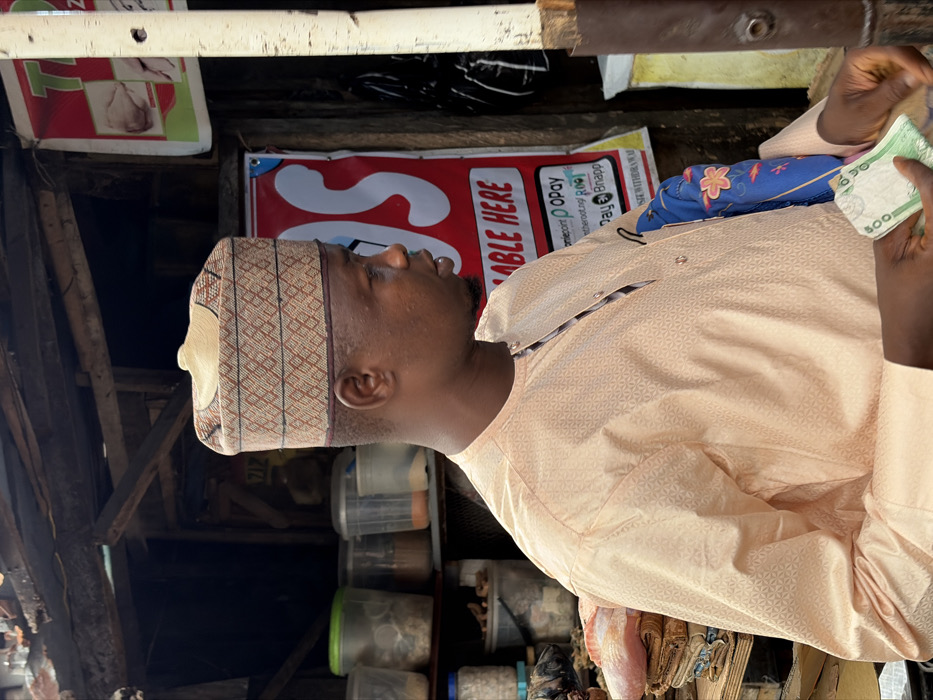 An adult male stands in profile, facing right, within a market stall in Jakande, Nigeria. He has a dark complexion and wears a light peach-colored, long-sleeved traditional garment with a subtle geometric pattern, and a tall, multi-patterned cap. His hands, in the lower frame, hold several Nigerian Naira banknotes, including 500 and 200 denominations. A blue object with pink floral designs is tucked under his right arm. The background reveals a rustic wooden stall structure with shelves holding various clear plastic containers of dry goods. A red banner displays payment service logos for "onepoint," "OPay," and "Pay Bsnapp" with the text "ABLE HERE." A partial sign above shows food items. A light-colored vertical post is visible on the far right.
