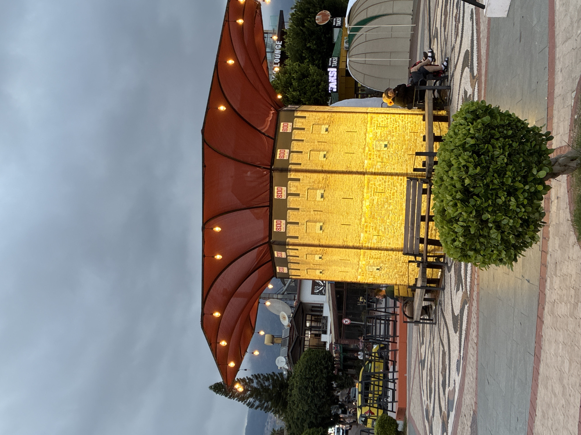 The image captures an outdoor commercial area at dusk or early evening in Alanya, Turkey. A brightly illuminated, yellow-toned building resembling a medieval fortress or tower dominates the center, featuring small, rectangular openings. Extending over an adjacent outdoor seating area is a large, reddish-brown fabric canopy adorned with numerous string lights.
Several tables and chairs are scattered across the paved ground, some occupied by individuals. One person in a yellow shirt is visible seated at a table near the illuminated building. A round, well-maintained green bush sits prominently in the foreground. In the background, other buildings, trees, satellite dishes, and distant mountains are discernible under a cloudy, overcast sky.
The activity suggests patrons are dining or socializing in an inviting atmosphere created by the warm lighting. Visible text on the canopy repeatedly shows "800". A sign in the background partially displays "LOUNGE" and clearly reads "TAKSİ," indicating a taxi service. The ground is paved with decorative, mosaic-like patterns alongside plain sections.