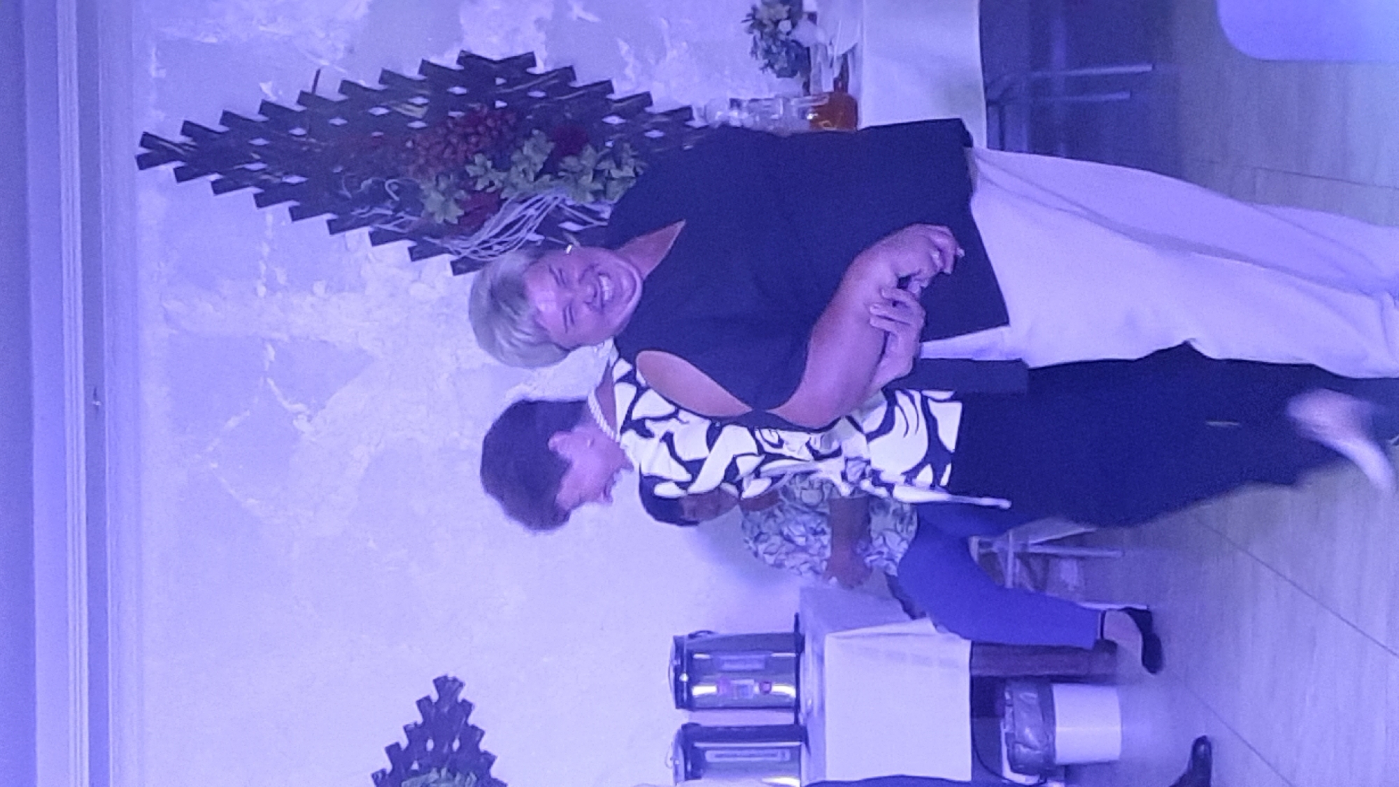 Two women are dancing at an event in Жодзішкаўскі сельскі Савет, Belarus. The woman on the right, with short blonde hair, is wearing a black off-the-shoulder top and cream-colored pants. She is laughing heartily. The woman on the left, with short brown hair, is wearing a white top with a black abstract pattern and black pants. She is also smiling. They are holding hands. In the background, there is a table with a trash can and two thermoses. Another person is seated in a chair in the middle ground. The environment is dimly lit with purple lighting.