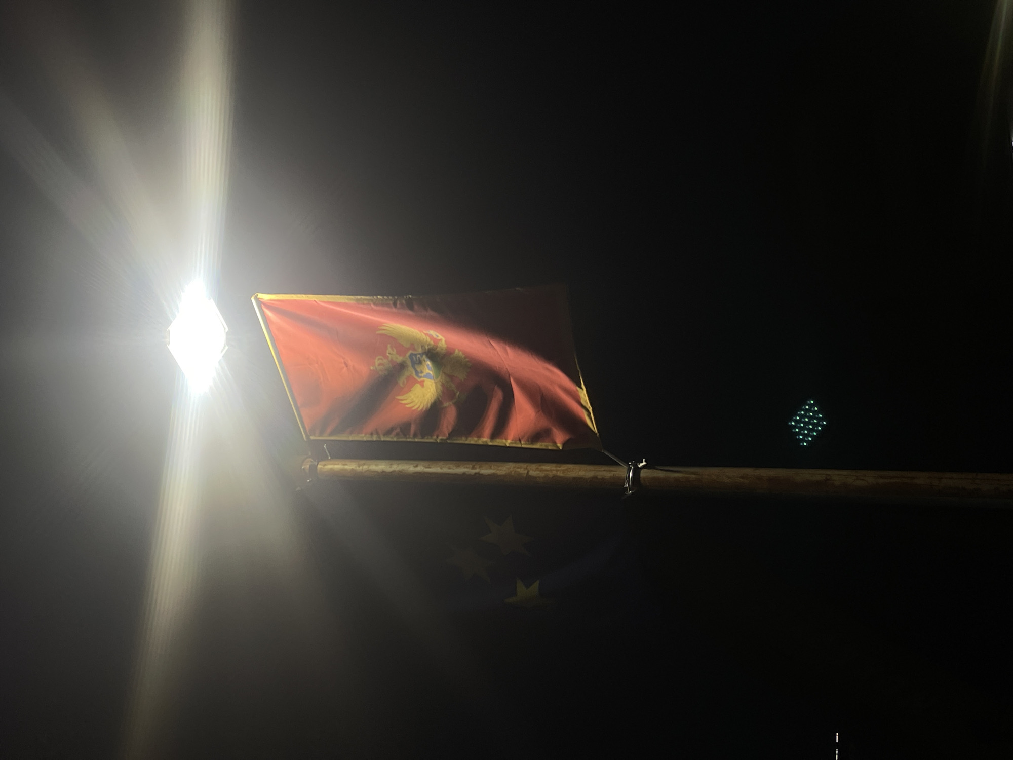 An image taken at night shows a flagpole with a flag of Montenegro hanging from it, illuminated by a bright overhead lamp. The flag is red with a golden double-headed eagle crest in the center. To the left of the flagpole, partially visible and appearing to be a separate flag or banner, are three yellow stars on a dark blue background. A traffic light with green lights arranged in a diamond pattern is visible in the lower-middle section of the image. The scene is dark, with the bright light from the lamp creating lens flare and a hazy effect. The background is black, suggesting the image was taken in an area with minimal ambient light.